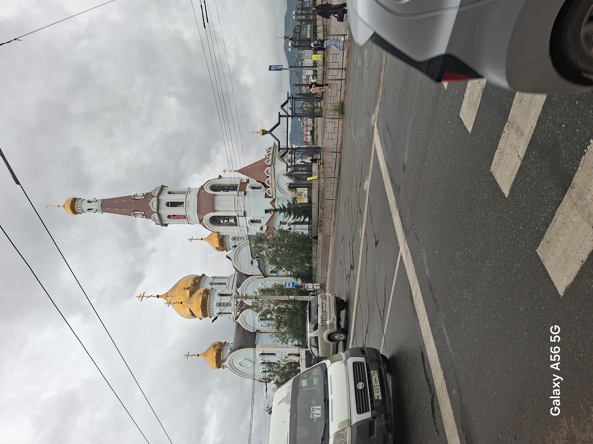 A frontal, slightly low-angle, medium shot captures a large, light blue Orthodox church with golden domes under a cloudy sky. The church has a prominent bell tower with a red roof. In the foreground, a white van with license plate K 455 XM 75 is on the left, facing right. To its right, a silver SUV is driving away. The scene is a street intersection with wet pavement, indicating recent rain. Several power lines are visible overhead. On the right side of the street, a black iron fence encloses the church grounds. People are visible walking near the entrance of the church. A silver car is partially visible in the lower right corner, also on the wet road. The bottom left corner of the image displays "Galaxy A56 5G".