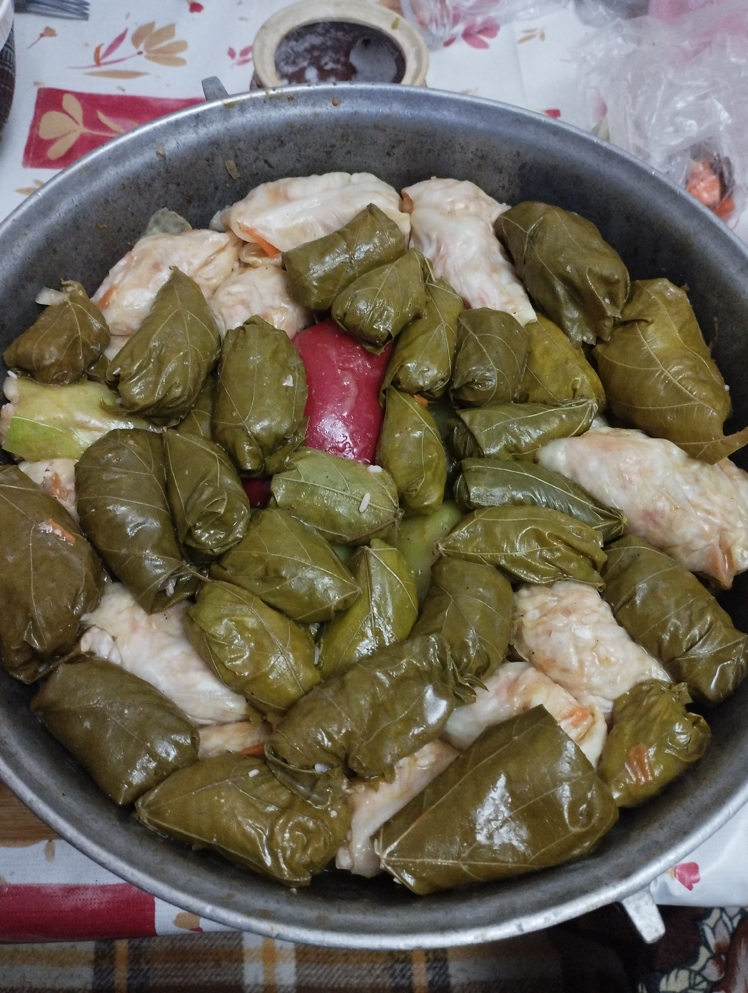 In the city of Fălești, Moldova, a close-up, overhead shot captures a large pot filled with dolma, a dish of stuffed grape leaves and cabbage rolls. The pot, appearing to be made of aluminum, is nearly full of the savory parcels. The dolma are a mixture of green grape leaves and white cabbage leaves, tightly rolled and packed together. Some of the cabbage rolls are visible at the bottom of the pot, with their white exterior peeking through. A few red items, possibly peppers or tomatoes, are interspersed among the dolma. The scene is set on a table covered with a tablecloth featuring a floral pattern in muted colors, accented by a red and white striped border. The lighting suggests an indoor setting, possibly a kitchen or dining area. There are no people visible in the frame, and the focus is solely on the food and its presentation within the pot.