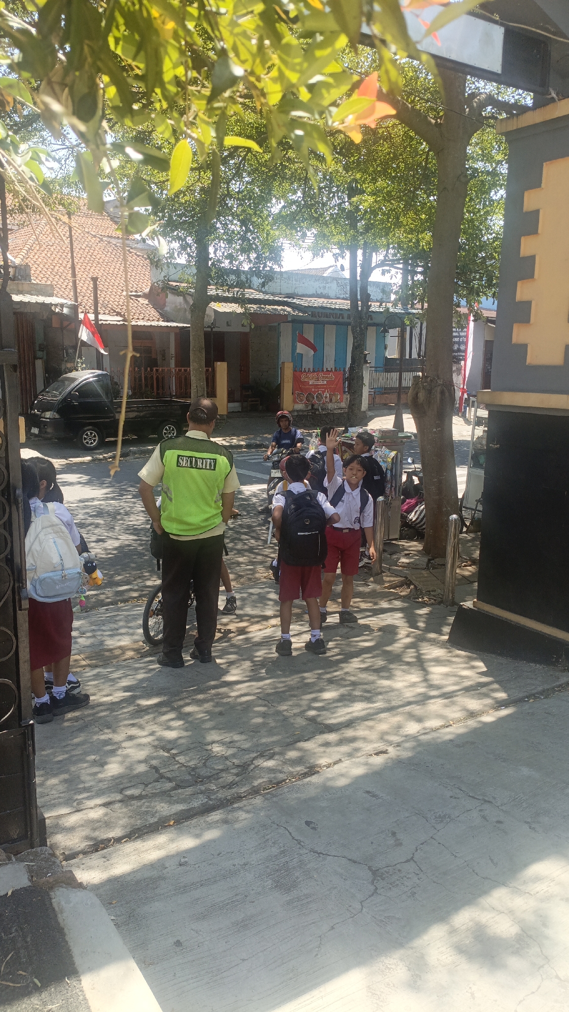 The image depicts a street scene in Salatiga, Indonesia, under a clear, sunny sky. In the foreground, a security guard wearing a bright green vest with "SECURITY" printed on it stands facing away from the camera. He is wearing dark pants and shoes, and a cap. To his left, two young girls in school uniforms, one with a light blue backpack and the other with a white backpack, are standing on the sidewalk.
Further down the street, a group of young boys in school uniforms are gathered. One boy is raising his hand, seemingly interacting with someone off-camera. He is wearing a white shirt, red shorts, and a black backpack. Another boy next to him is also wearing a white shirt and red shorts, with a black backpack. A third boy in similar attire is partially visible behind them.
Several motorcycles are parked or being ridden in the background. A black pickup truck is parked on the left side of the street. Buildings with red-tiled roofs and colorful facades line the street. Indonesian flags are visible, attached to some of the buildings, indicating a possible national celebration. The ground is paved with concrete, and dappled sunlight filters through the trees overhead, creating a pattern of light and shadow. The overall atmosphere appears to be one of everyday life, possibly during school dismissal or arrival.