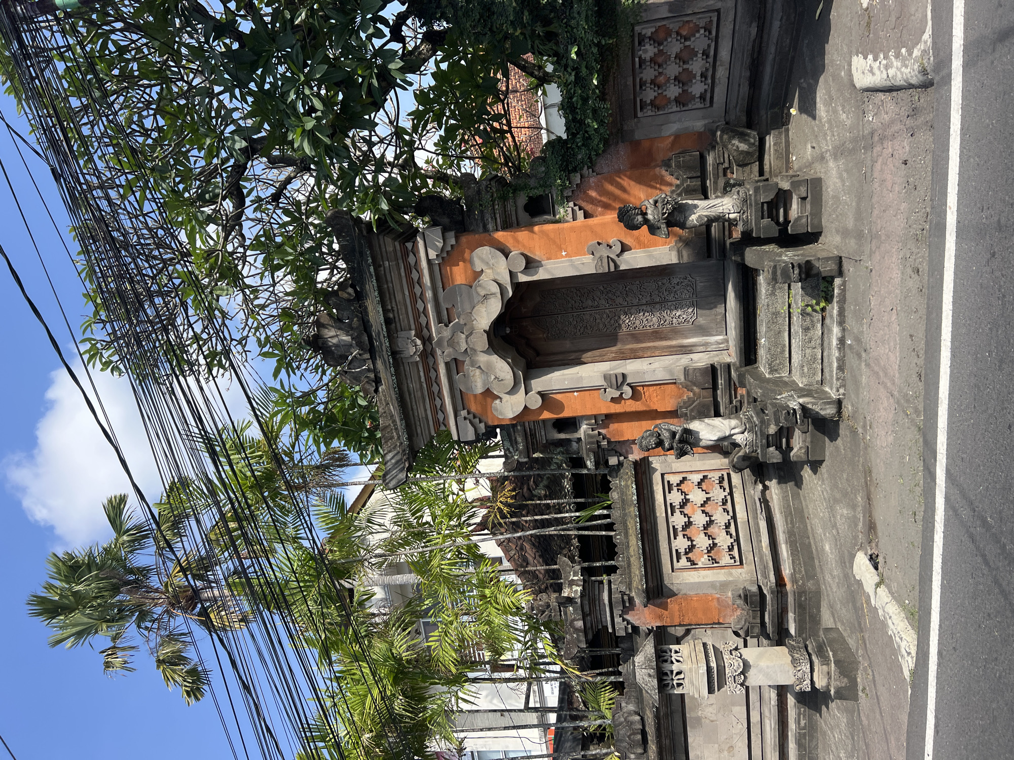 This image captures a street view of a traditional Balinese entrance gate in Denpasar, Indonesia. The gate is ornate, constructed with dark wood carved with intricate patterns, and framed by reddish-orange stone walls. Two stone statues of Balinese figures stand guard at the entrance steps. The gate is surrounded by lush tropical foliage, including palm trees and other greenery, which create a sense of seclusion. Overhead, a dense network of black electrical wires crisscrosses the bright blue sky, punctuated by scattered white clouds. The scene is viewed from a street level, with a paved road and a white line marking the edge in the foreground. The architecture suggests a residential or temple property, typical of Balinese design.