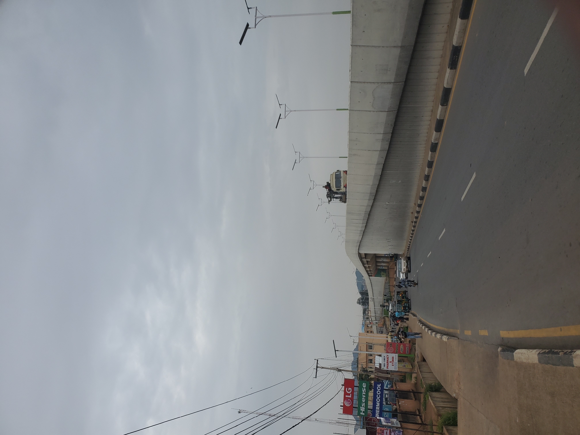 This image depicts a street scene in Jos, Nigeria. The foreground shows a paved road with a yellow and black striped curb, leading towards a concrete overpass or flyover. On the left side of the road, there is a sidewalk and a row of shops with various signs, including "LG", "Hisense", and "THERMOCOOL". Electrical wires hang overhead, supported by utility poles.
The overpass is a prominent feature, made of concrete with a textured surface. Several streetlights with solar panels are installed along the top of the overpass. In the distance, more streetlights are visible, as well as buildings and hills.
The road itself has a few vehicles, including cars, a motorcycle, and a tricycle (popularly known as a "keke"). Several people are walking and standing around, some near the shops and others on the road. The sky is overcast and gray, suggesting a cloudy or potentially rainy day. The overall impression is a busy urban environment undergoing some infrastructure development, evidenced by the overpass and new streetlights.
