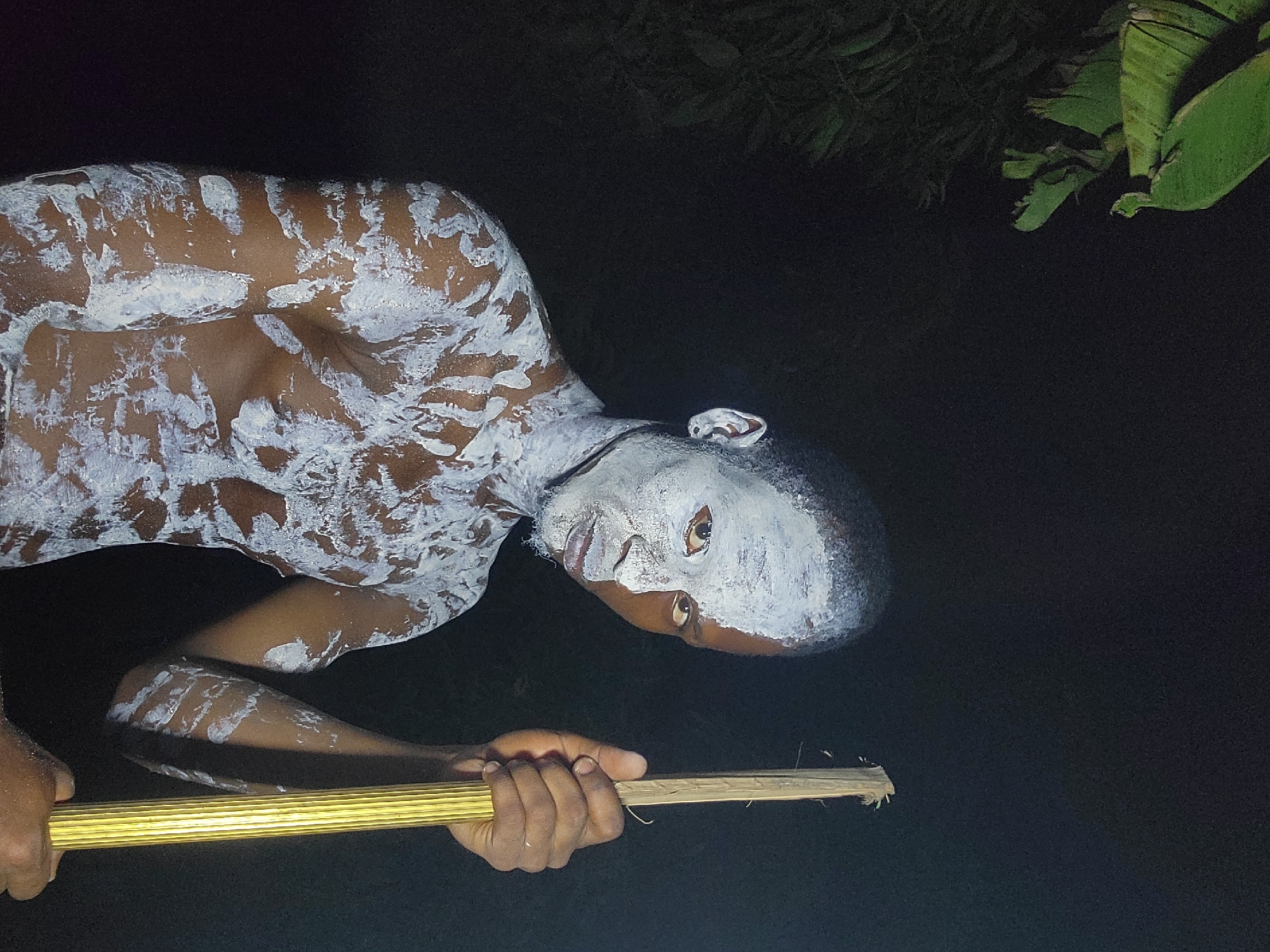 A dark-skinned person with white paint covering their body and face is holding a bamboo-like rod. The background is dark, with foliage visible in the top right corner. The location is likely Onitsha, Nigeria, based on the cultural associations with body paint and bamboo. The person is angled towards the camera.