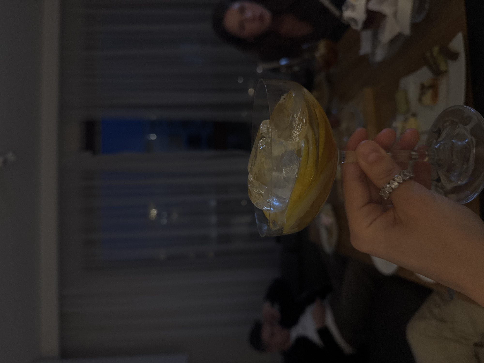 A person is holding a wine glass filled with ice and a lemon wedge. The person is wearing a ring. In the background are other people and food on a table. The location is possibly in a restaurant or bar in Warsaw, Poland, given the urban background visible through the window.