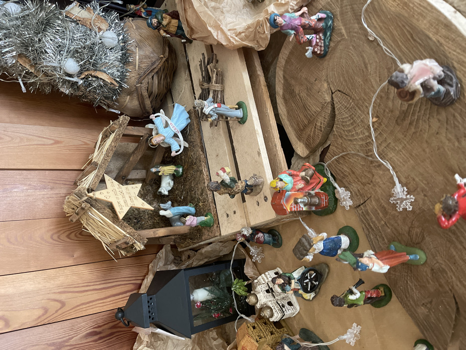 This media file captures a detailed, rustic Christmas nativity scene, known as a "presepio," likely set up indoors in Castione, Italy. The scene is composed of numerous small, hand-painted figurines arranged on various wooden surfaces, including wooden planks, a large cross-section of wood with visible rings, and a wooden floor.
The central part of the display features a small, simple stable or manger crafted from twigs and straw. Above it hangs a light-colored wooden star with an inscription in Italian: "Ti appeso io penso a te come il cielo mi accende la stella," which translates to "I hang you, I think of you as the sky lights up the star."
Numerous miniature figures, possibly "Santon" style, populate the scene. These include an angel with a banner reading "GLORIA," various villagers and shepherds in traditional attire, some carrying bundles of sticks, others kneeling or standing. A figure resembling a woman in blue, perhaps Mary, and another figure on a donkey are also visible. One distinct figure is clad in red with what appears to be a chimney or oven, suggesting a baker.
Other decorative elements include a silver tinsel decoration, possibly a small Christmas tree or wreath, adorned with white baubles and tiny lights, positioned to the left. String lights with snowflake-shaped covers are woven throughout the display, adding a festive glow. A black, traditional-style lantern containing a white candle and sprigs of greenery with red berries sits on a lower level, alongside other figures and brown crumpled paper used as ground cover or filler.
The overall arrangement gives the impression of a vibrant and traditional holiday display, evoking a sense of warmth and festive spirit. The collection of diverse figures and rustic elements points to a cherished seasonal tradition.