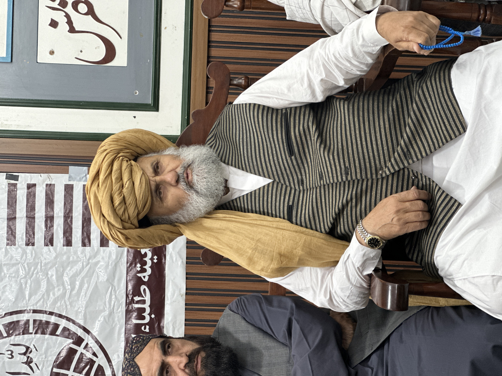 An elderly man with a turban and a grey beard is seated, holding blue prayer beads. He is wearing a striped vest and a white shirt. A watch is visible on his left wrist. Another person is partially visible below him. The setting appears to be indoors, with patterned wooden walls and a banner partially visible in the background. The banner has Arabic script and a symbol on it. This is likely a scene somewhere in the city of Quetta City Tehsil, Pakistan.