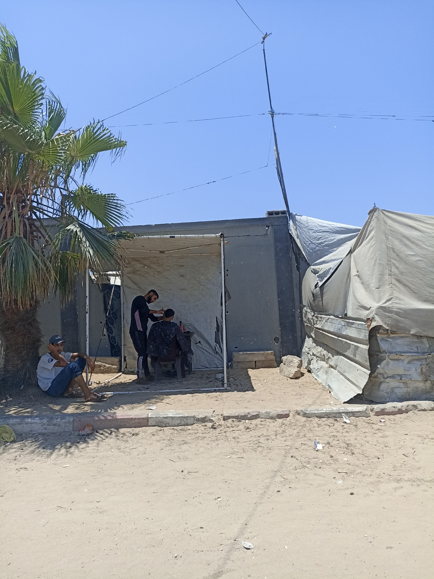 In the city of Gaza, Palestinian Territories, on a sunny day with a clear blue sky, two men are having their hair cut under a makeshift canopy. One man, wearing a black shirt with pink stripes and black pants, stands and operates clippers on the head of another man, who is seated and wearing a dark, patterned shirt. A third man, wearing a gray t-shirt and blue jeans, sits on the ground to the left, leaning against a palm tree and looking away from the haircut. The scene is set in a sandy area with a rough, unpaved ground. To the right of the haircutting station, a larger tent made of canvas is erected. The structures appear to be temporary and constructed from various salvaged materials, including corrugated metal sheeting. The overall environment suggests a context of improvisation and resourcefulness.