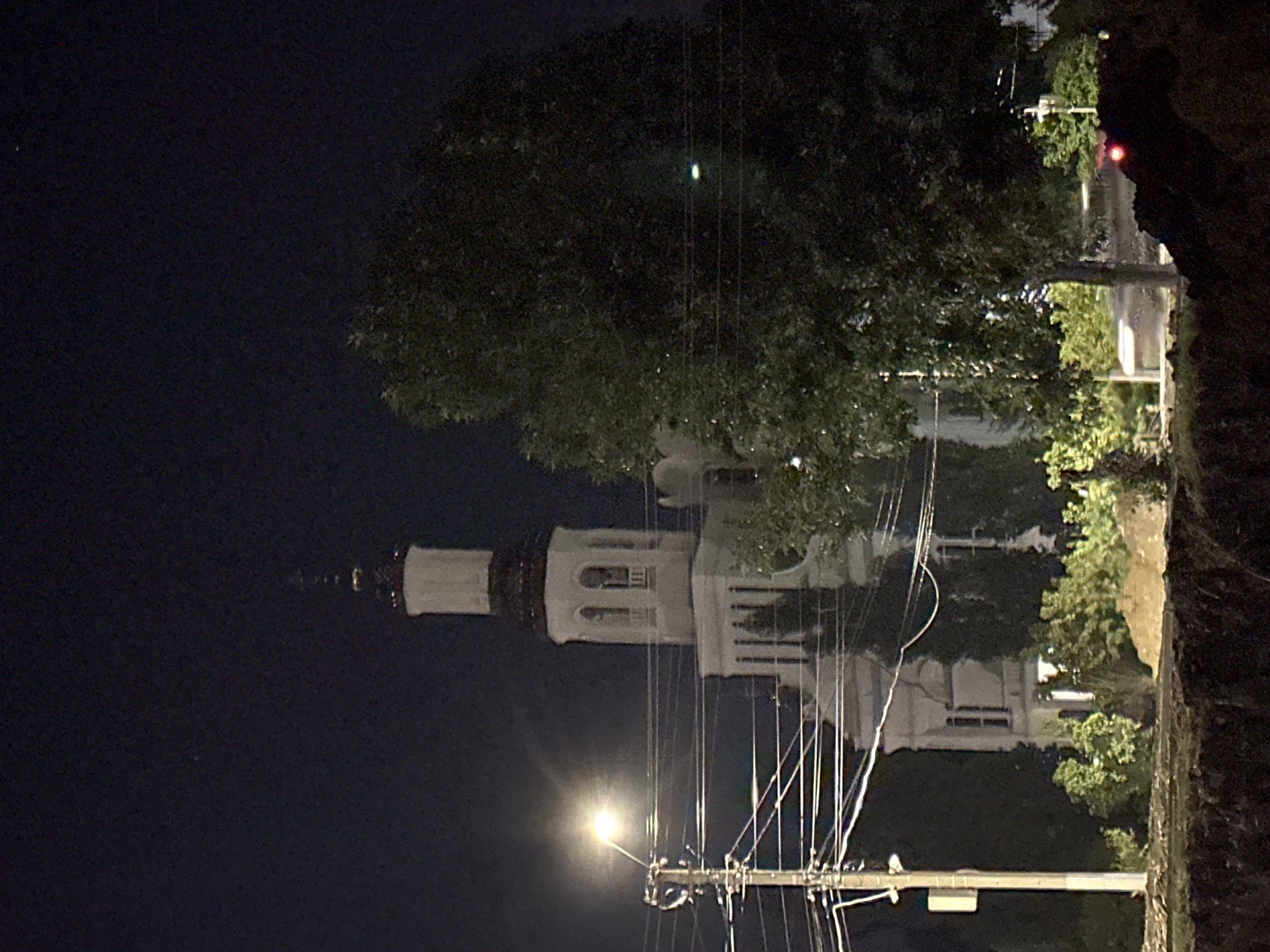 The image shows a nighttime view of a white building with a tall, multi-tiered tower, possibly a church or a campanile, in Nevinnomyssk, Russia. The building features arched windows and a dark, conical roof on the tower. In the foreground, a power pole with a bright streetlamp is visible, along with numerous power lines crisscrossing the frame. A large, dark tree dominates the right side of the image, partially obscuring the building and the sky. The ground in the immediate foreground is dark and appears to be uneven, possibly indicating construction or rough terrain. The sky is dark and clear, with a few faint stars visible. The overall lighting is dim, with the streetlamp providing a strong, localized light source.