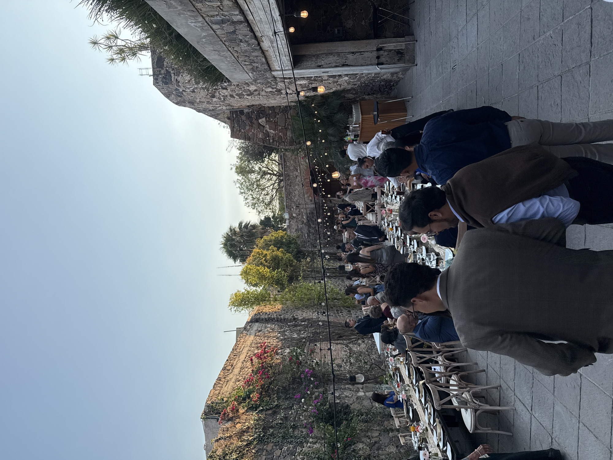 The image depicts an outdoor event with numerous people seated at a long table set for dining. The location appears to be in Santiago de Querétaro, Mexico, given the architectural style featuring stone walls and colonial-era buildings in the background. People are dressed in formal attire. String lights are strung overhead, illuminating the scene. The event looks like a wedding reception or a formal dinner. The foreground shows a close-up of the guests, and the background shows a long line of seated people. The environment consists of a stone patio and historic stone walls.