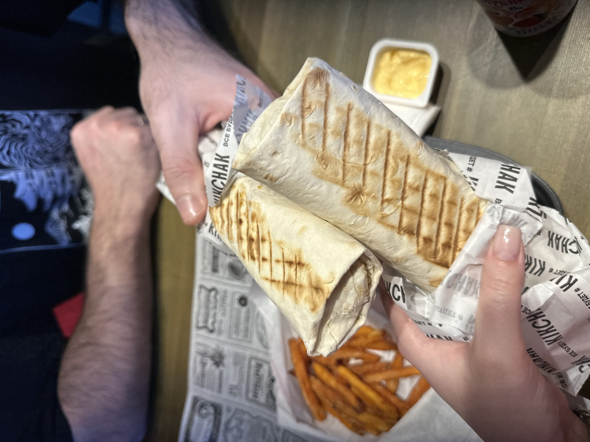The scene depicts an interior dining setting in Rostov-on-Don, Russia, focusing on a casual meal shared by two individuals. In the foreground, a person with light-toned skin and clear-polished fingernails holds two large, grilled wraps, oriented diagonally towards the upper right. These wraps are encased in white paper featuring black text, prominently displaying "КИКЧАК" (KIKCHAK) and "ВСЕ БУДЕТ" (VSE BUDET). To the left, a white paper container holds a quantity of orange-hued french fries, likely sweet potato fries. In the background, the forearms and hands of a second individual are visible, resting on a light brown wooden or laminate table. This person's left hand is near a crumpled piece of paper with similar branding, while their right arm extends towards the right edge of the frame. The individual wears a dark long-sleeved garment with a subtle textured pattern. To the right of the wraps, a small white rectangular dish contains a viscous yellow condiment. A portion of a cylindrical object, possibly a drink container with an orange label, casts a shadow on the far right. The overall lighting is subdued, suggesting an indoor restaurant or cafe environment.