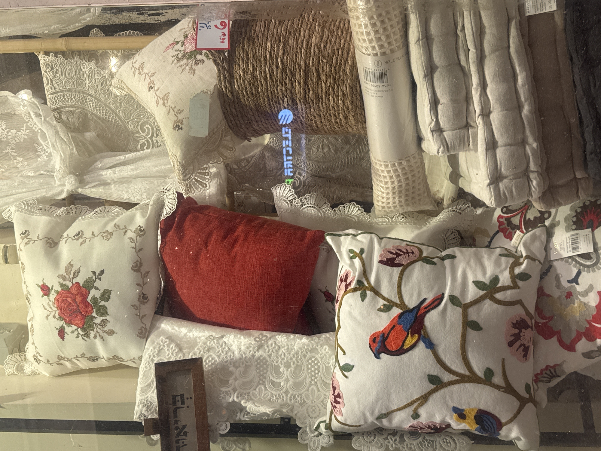 The image shows a display of textiles behind glass, likely in a shop in Ramat Gan, Israel. Featured are decorative pillows: one with a red rose design, another with an embroidered bird and floral motif, and a red cushion. There are also rolls of material, possibly burlap and quilted fabric, along with lace and other fabrics. A price tag in Hebrew is visible. The scene is a close-up view of a retail display.