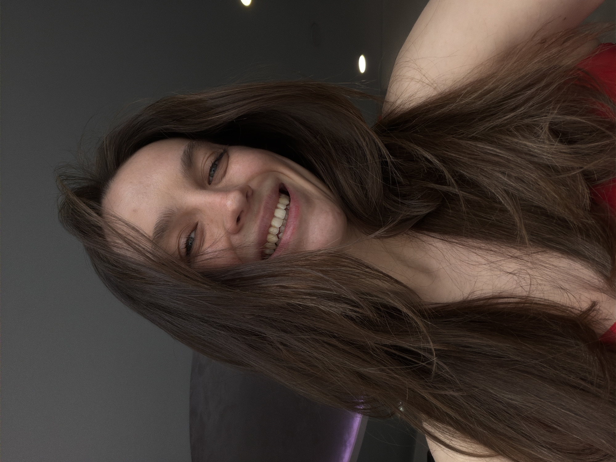 An indoor close-up portrait of an adult female in Budva, Montenegro, depicts her smiling broadly directly at the camera. She has fair skin, blue eyes, and long, layered brown hair that frames her face and extends over her shoulders and upper chest. Her smile reveals both upper and lower teeth. A red garment is visible on her lower chest area. The background is a dark grey wall or ceiling, with a dark grey ceiling panel visible on the upper left. Two distinct circular recessed lights, emitting warm white illumination, are visible in the upper right quadrant of the background. A subtle linear purple light accent is visible on the far left, suggesting architectural lighting. The image appears to be taken from a slightly low angle.