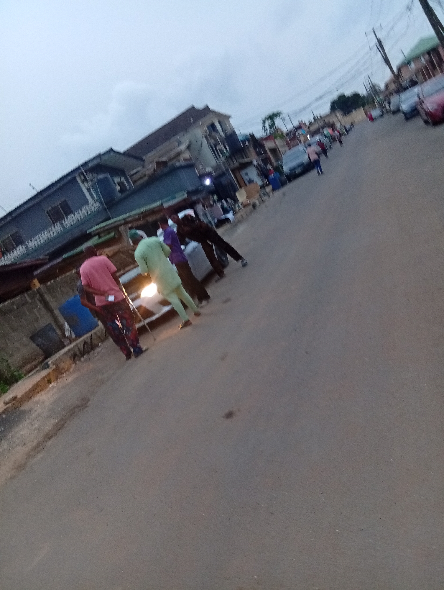 This image captures an urban street scene in Shangisha, Nigeria, likely during dusk or dawn under an overcast sky, as indicated by the dim natural light and the illuminated headlights of a car. The photograph is taken from a slightly tilted perspective and appears somewhat blurry.
In the foreground, a group of men are standing on the asphalt road. One man in a pink shirt and patterned trousers is closest to the camera, while another in a light green outfit stands near a white car with its headlights on. A third man wears a purple shirt. Their exact activity is unclear, but they appear to be gathered or conversing around the vehicle. A blue barrel is visible on the roadside near the foreground figures.
Further down the street, more individuals are seen walking or standing, and several cars are parked along both sides of the road. The street is lined with multi-story buildings, some featuring balconies and fences. Utility poles and a network of power lines crisscross the sky above the buildings. The road is paved, with rougher, unpaved sections and some debris along its edges. No discernible text is visible anywhere in the image.
