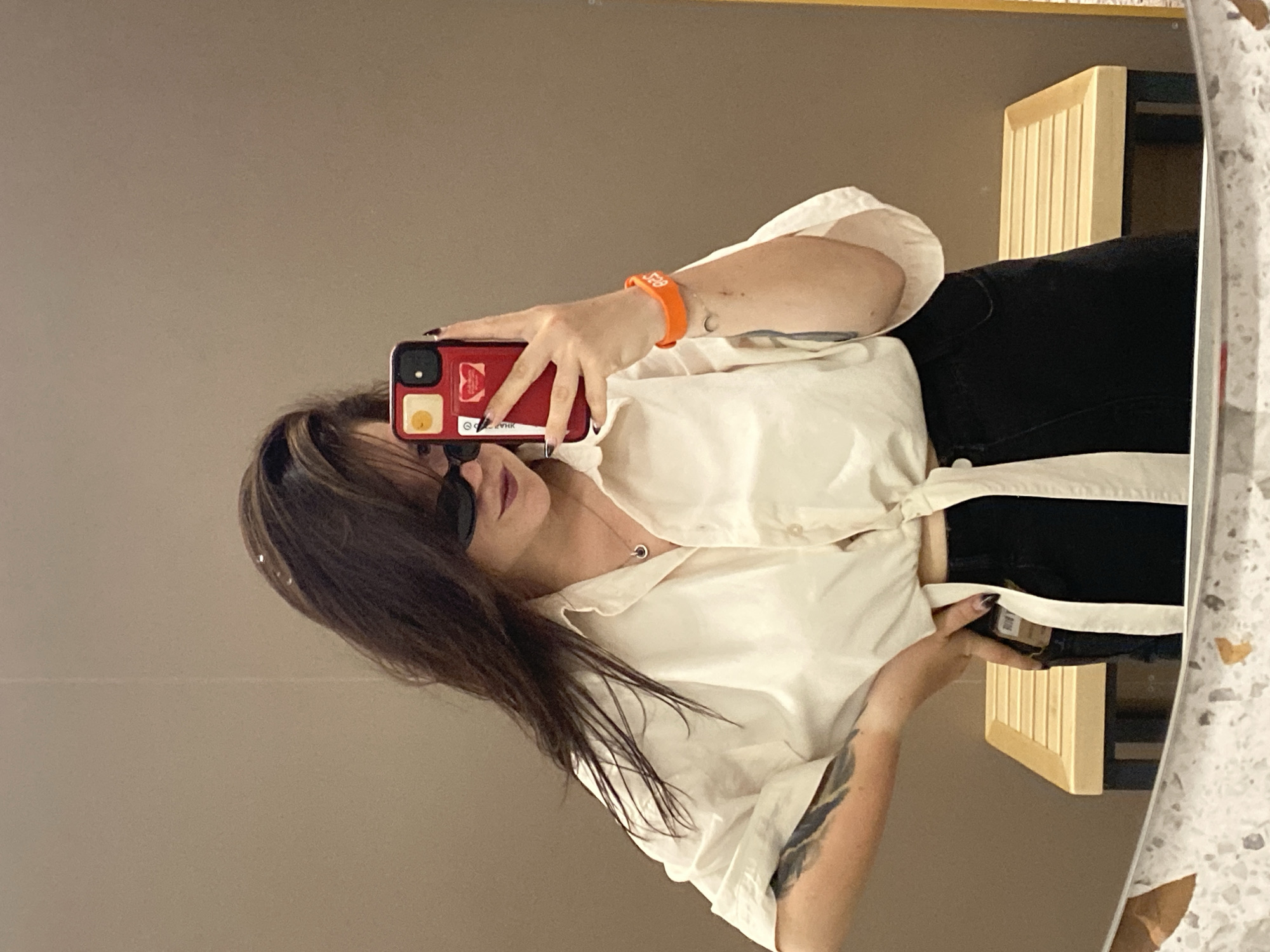 A young woman is taking a mirror selfie in what appears to be a changing room or dressing area. She has long, dark hair, and is wearing oversized sunglasses, a white button-up shirt tied at the waist, and black pants. She is holding up a red phone in her right hand, which is decorated with stickers. She is also wearing an orange wristband on her right arm and a delicate necklace. A tattoo is visible on her left arm. In the background, a wooden bench with slats is visible. The walls are a neutral beige color. The mirror reflects the room and the woman. The image is not specific to Votkinsk, Russia.