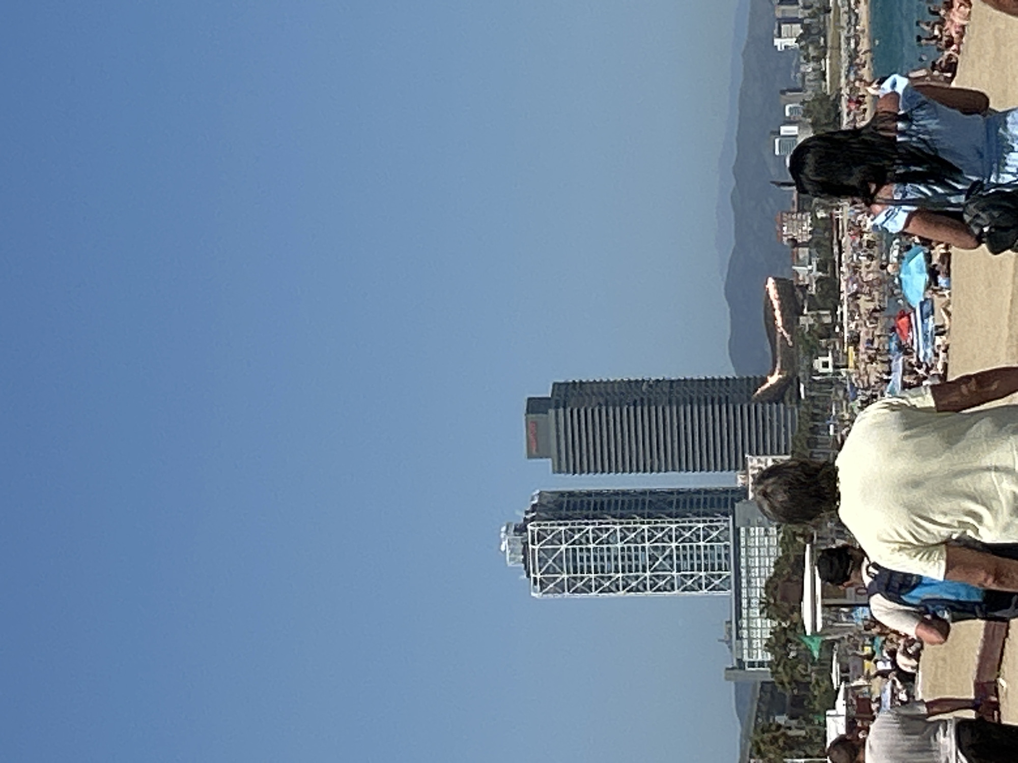 The image captures a sun-drenched beach scene in Barcelona, Spain, with a clear blue sky dominating the upper portion. In the mid-ground, two prominent skyscrapers stand against the horizon: one with a distinctive white geometric lattice exterior and another taller, darker building with horizontal louvers. A body of water, likely the Mediterranean Sea, is visible beyond the beach, dotted with numerous beachgoers and colorful umbrellas. In the foreground, several individuals are walking on the sand. A man in a yellow t-shirt is walking away from the camera, while a woman in a blue off-the-shoulder dress is also seen from behind. The beach is crowded with people sunbathing and enjoying the seaside. In the far distance, mountains form a hazy backdrop to the cityscape. A fiery object, possibly a flare or a jet trail, is visible in the sky between the buildings and the mountains.