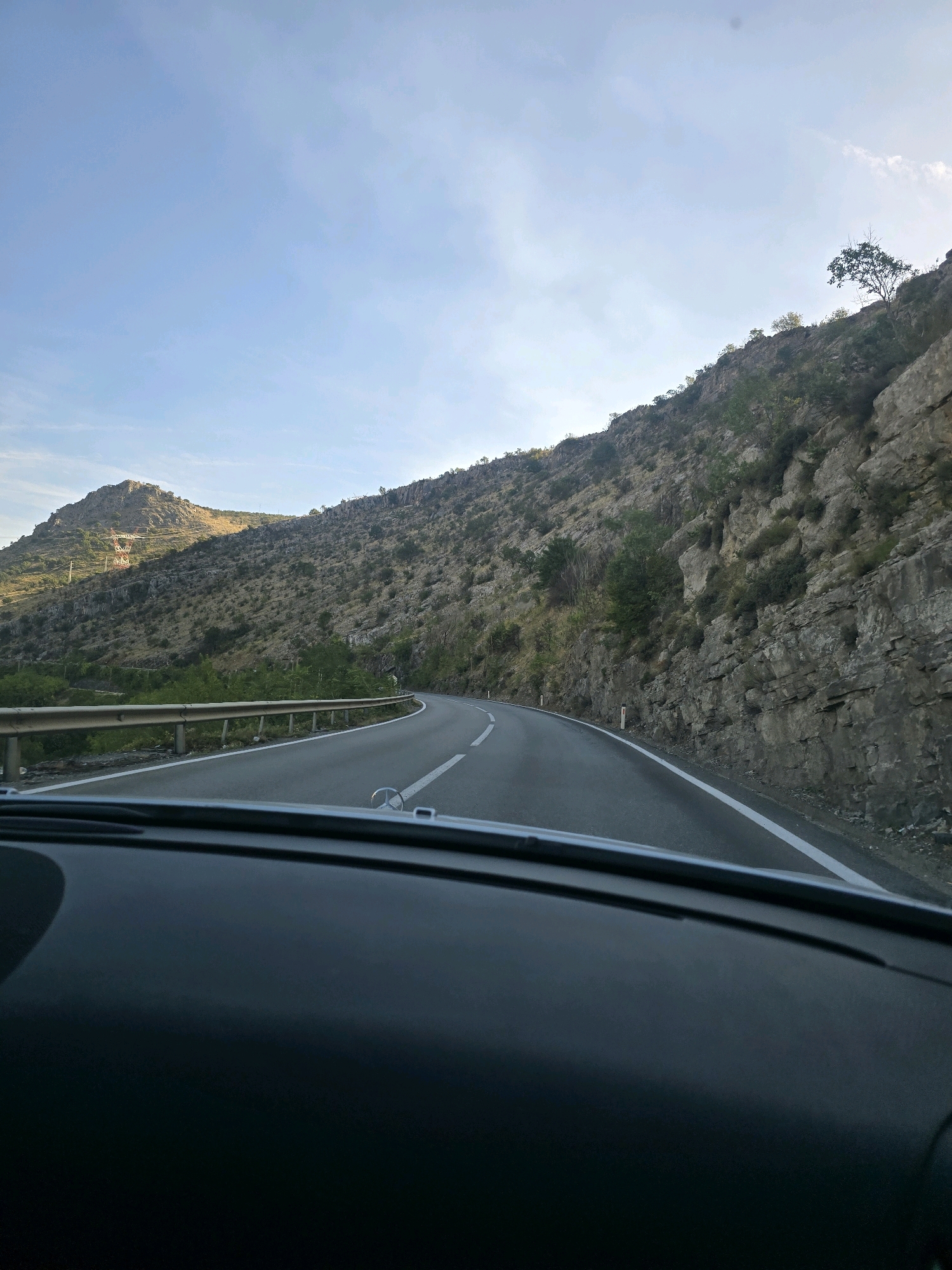 The image shows a view from a car dashboard looking out at a winding road through a mountainous landscape. The road curves to the left, bordered by a metal guardrail on the left and a steep, rocky cliff face on the right. The mountains are covered in sparse vegetation and large rock formations. In the distance, on the left side of the road, there are hills with some structures, possibly telecommunication towers. The sky is clear blue with some wispy clouds. The lighting suggests it is either early morning or late afternoon, with the sun casting a warm glow on the distant hills. The scene appears to be a scenic drive through a rugged, natural environment. Without additional context, it is not possible to pinpoint a specific location within Podgorica, Montenegro, as the image depicts a general mountainous road.
