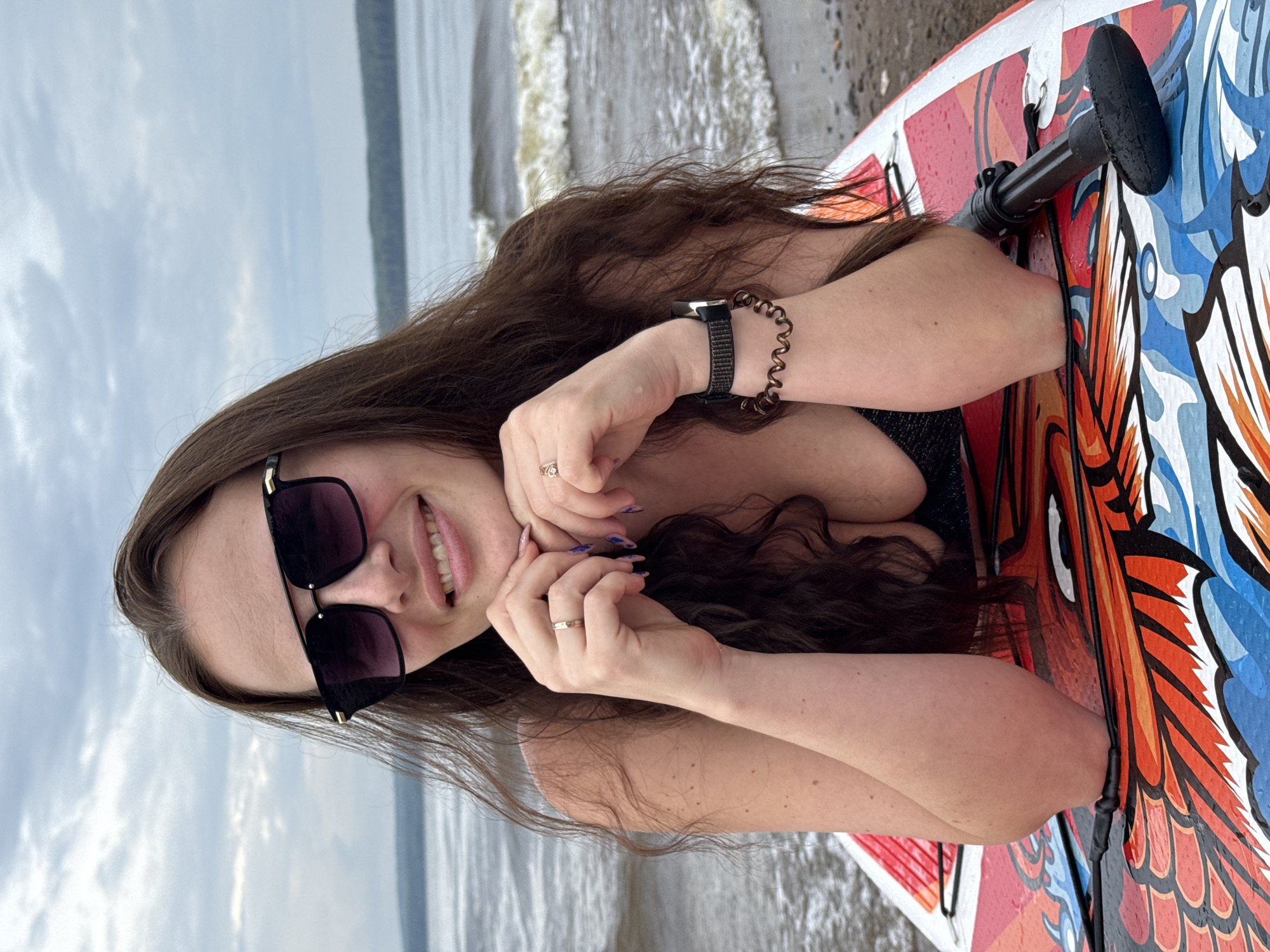 A woman with long, dark hair and sunglasses is lying on a colorful paddleboard on the shore of a body of water. The paddleboard has a design featuring a dragon motif with red, orange, and blue colors. The woman is wearing a black swimsuit. She is resting her chin on her clasped hands, which are adorned with rings. She is also wearing a watch and a spiral-shaped bracelet on her left wrist. The background shows a cloudy sky and a body of water with small waves. This scene is consistent with the Kama River embankment in Chaikovsky, Russia, a popular location for water activities and recreation.