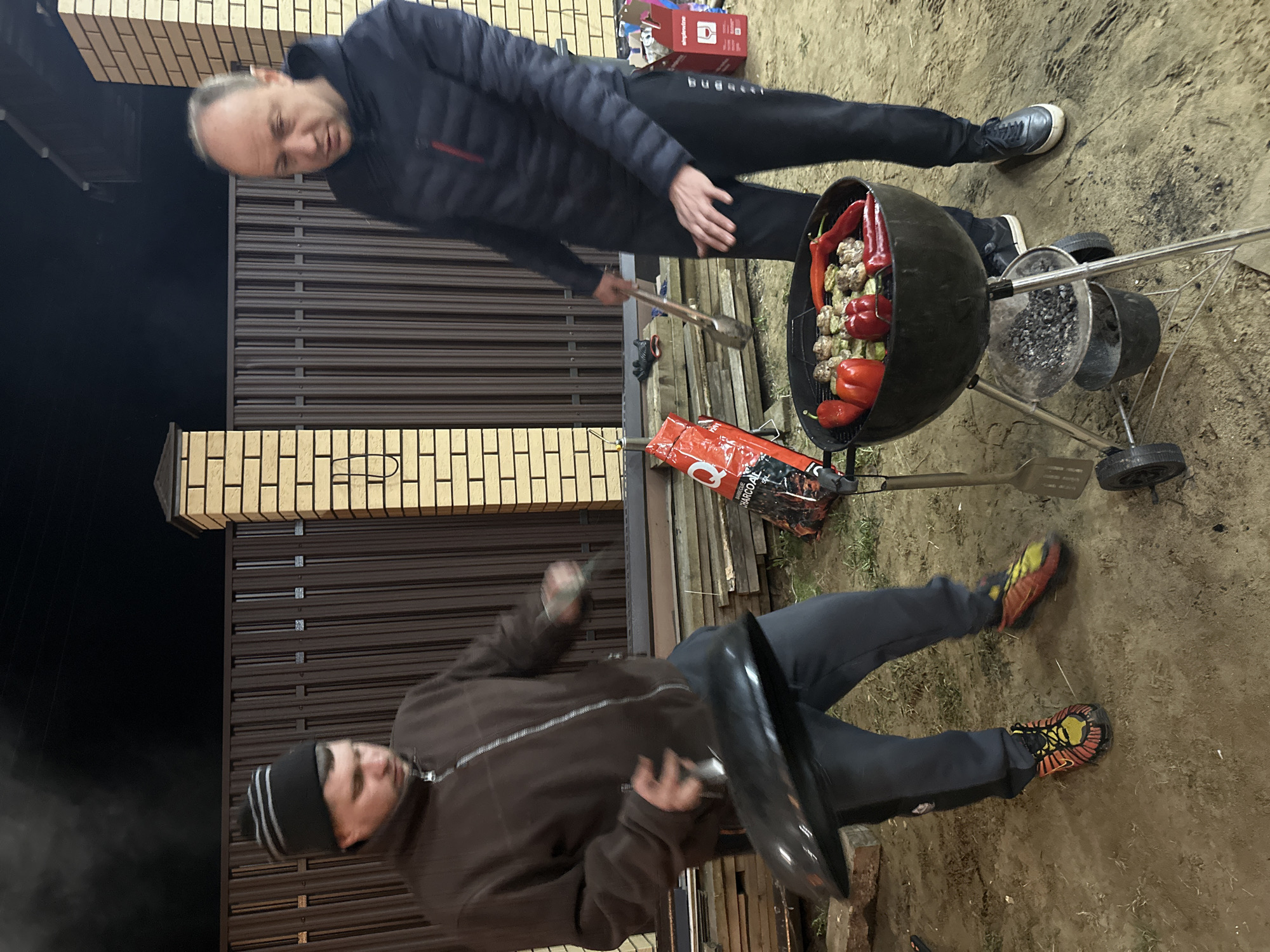A nighttime scene in слобода Старая, Russia, shows two men standing near a charcoal grill. One man, wearing a dark jacket and dark pants, stands over the grill holding tongs. The grill is filled with food, including red peppers and what appear to be mushrooms or meatballs. The other man, wearing a brown jacket, dark pants, and a beanie, is bending over and holding the lid of the grill in his hand. Bags of charcoal and cooking utensils are visible near the grill. The setting appears to be a backyard or outdoor space near a brown metal fence and a brick structure.
