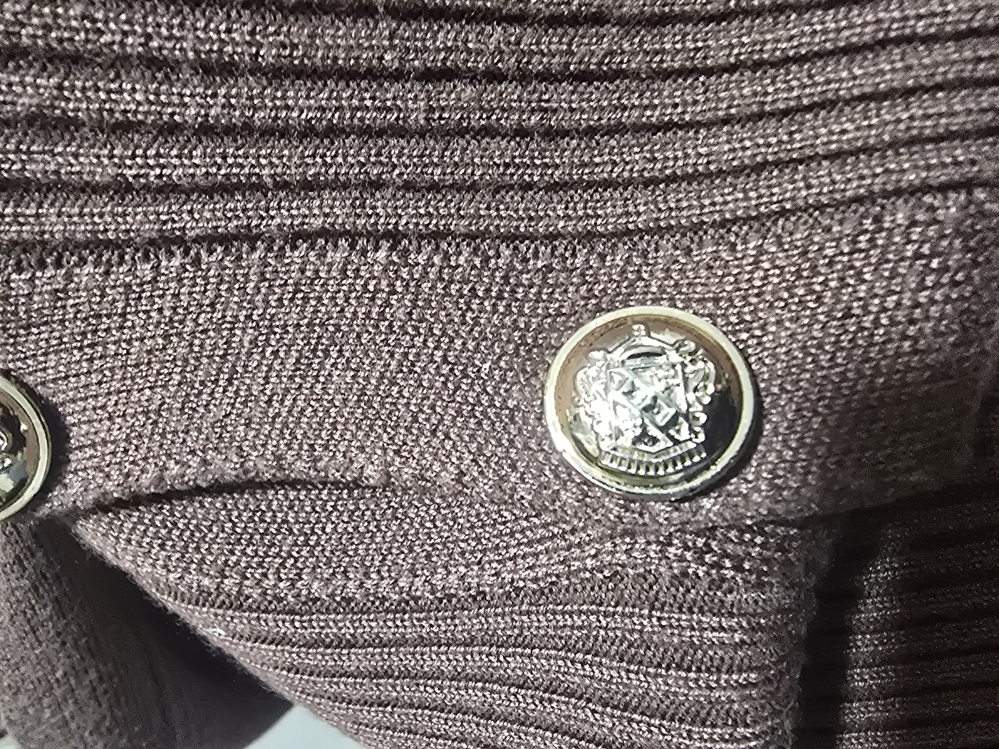 The image is a close-up shot of a brown ribbed knit fabric with two ornamental buttons. The button in the foreground is silver-toned and features a detailed embossed design. The fabric has a slight sheen, possibly due to the material composition. The image does not contain any contextual information about its location in Ulan-Ude, Russia, or any people or actions. It is a still life focusing on the texture and details of the garment.