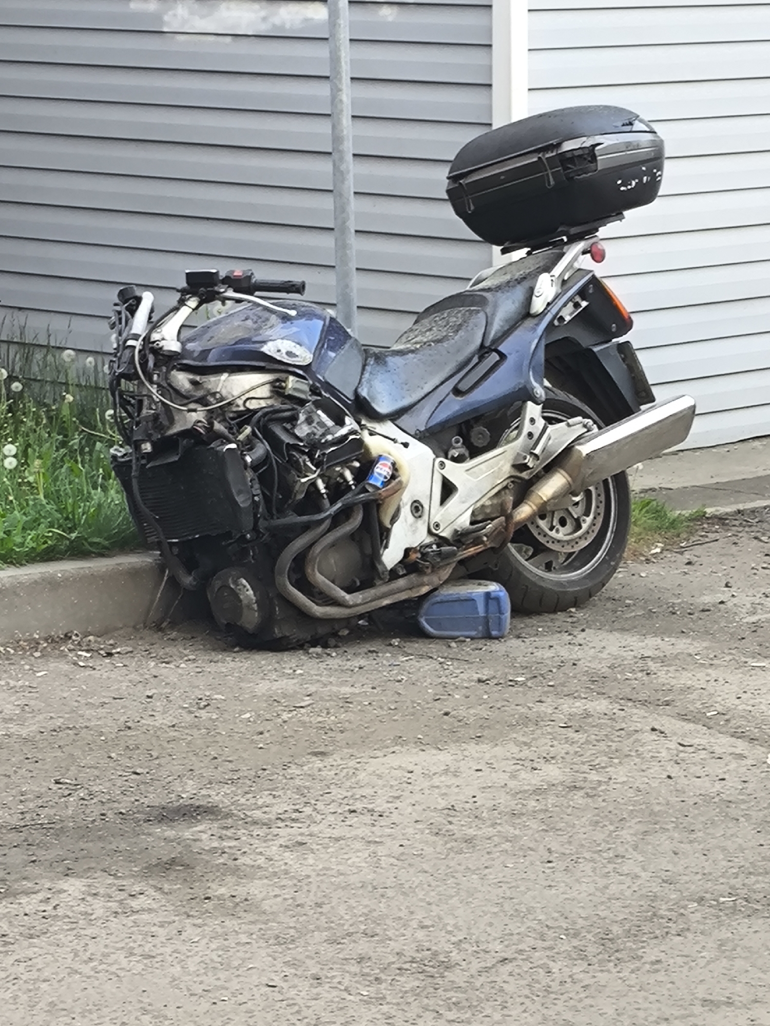 The image shows a blue motorcycle that appears to have crashed, resting against a curb. Its front is heavily damaged, with visible engine components and exposed wires. The motorcycle has a black storage box mounted on the rear. It is leaning against a curb with green plants growing behind it. A blue container supports the rear wheel. The motorcycle is positioned on a dirt or gravel surface next to a building with gray siding. A pole is situated behind the motorcycle. The location is a street in a city.