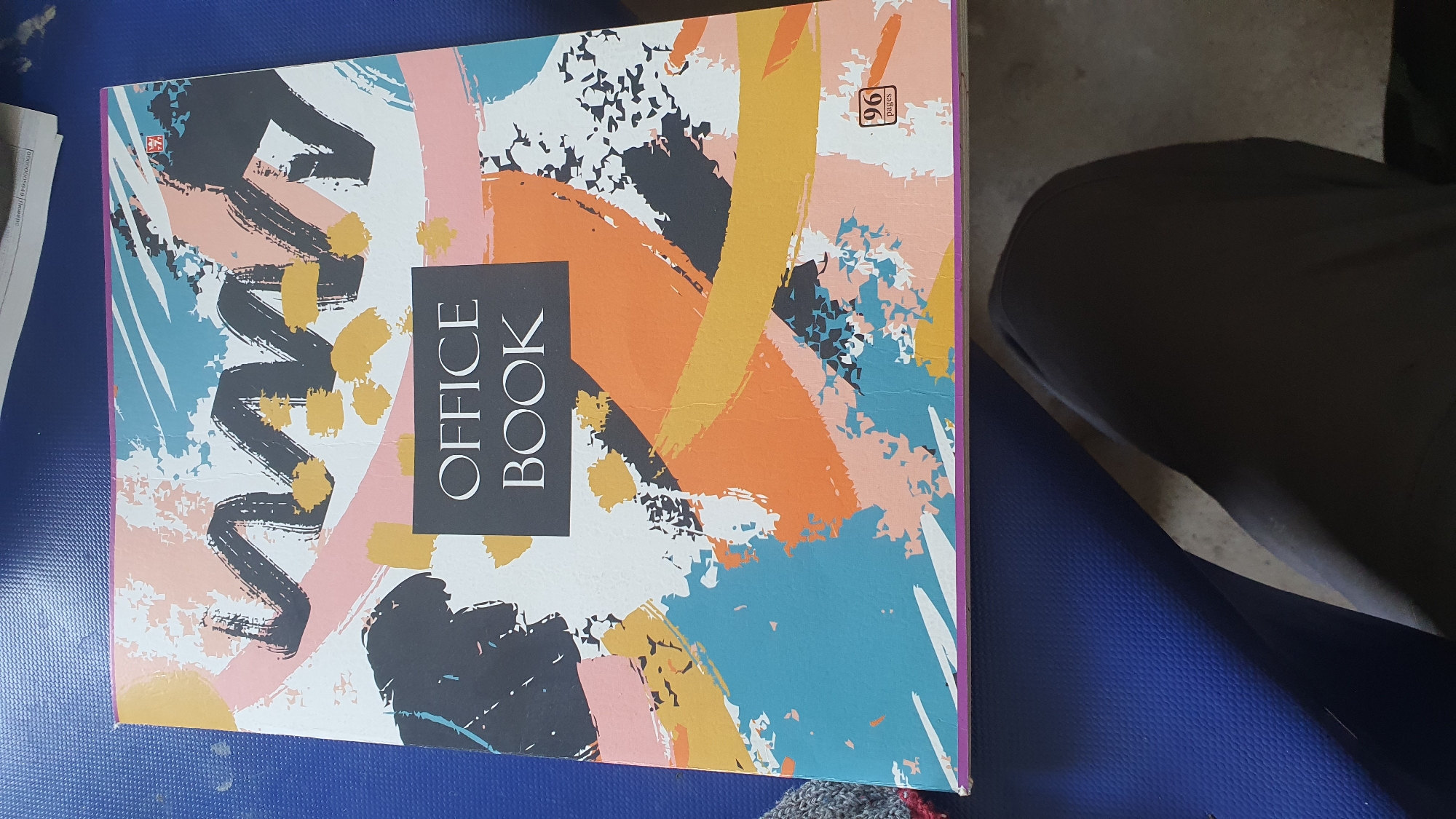 A close-up, top-down shot shows the cover of an "OFFICE BOOK" notebook, which has a colorful, abstract, brushstroke design. The notebook is lying on a textured blue surface. A portion of dark gray pants is visible in the lower right corner of the frame, suggesting a person is sitting or kneeling. The background is out of focus.