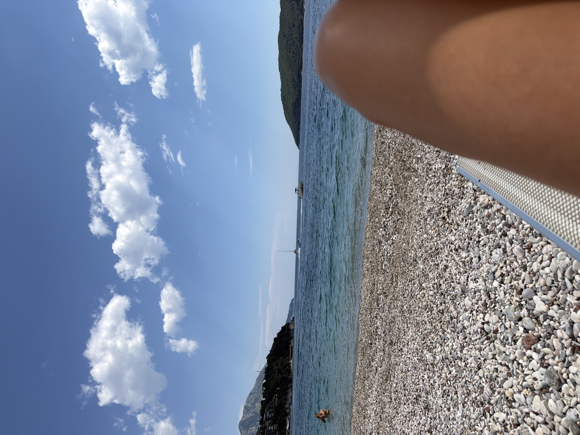The image shows a beach in Budva, Montenegro, on a sunny day with a blue sky and scattered white clouds. The beach is composed of small, rounded, grey and white pebbles. The Adriatic Sea is light blue-green, with gentle waves. Two sailboats are visible on the horizon. On the left side, there is a forested mountain range. A person is standing waist-deep in the water. Part of a person's leg and a metallic beach chair are visible in the foreground on the right side.