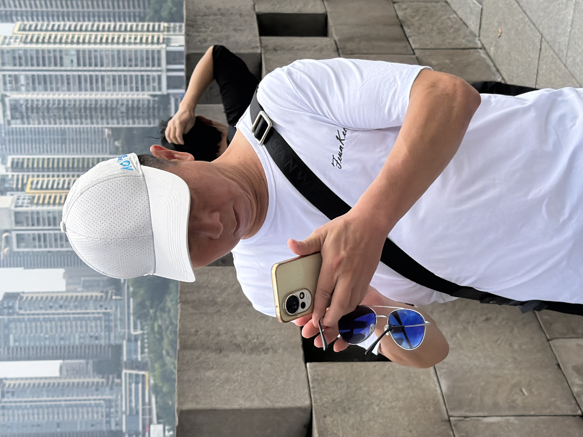A middle-aged East Asian man wearing a white t-shirt, white baseball cap, and a black crossbody strap is holding a gold smartphone and a pair of blue-tinted sunglasses in his hands. He is looking down at the phone, his brow furrowed. Behind him, a stone wall forms a barrier, and in the background, a dense cityscape of tall buildings is visible, suggesting an urban environment. The scene appears to be outdoors, possibly a viewing area or promenade in the Yuzhong District, China, given the architectural style of the surrounding buildings. Another person is partially visible in the background, also facing away from the camera.