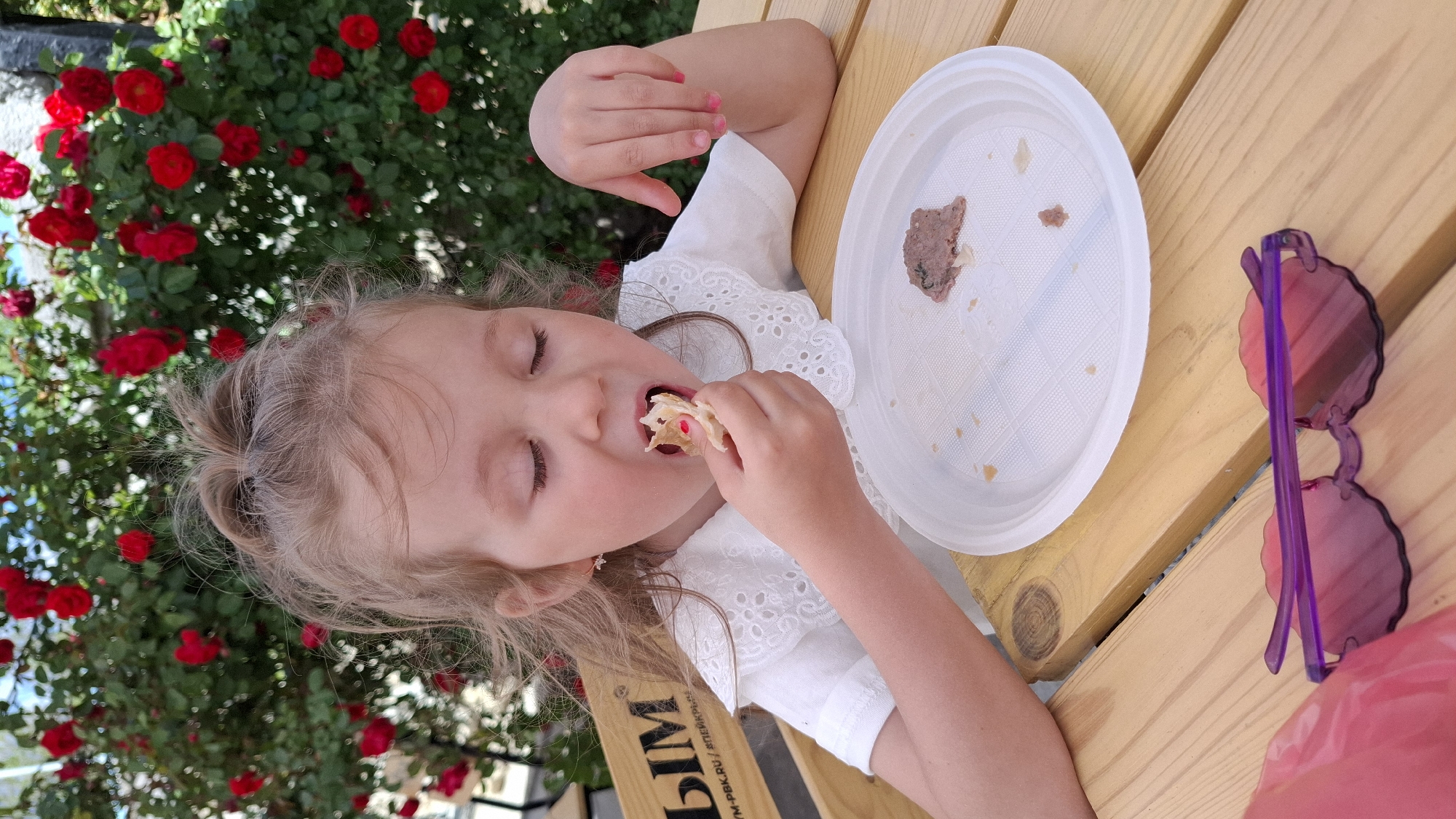 A young girl with blonde hair, wearing a white dress, is seated at a wooden table outdoors, with a plate of food in front of her. She is eating a small piece of food with her eyes closed. On the plate, there are remnants of food, suggesting she has already eaten some of it. To the right of the plate, there is a pair of pink sunglasses. In the background, there is a bush of red flowers. The location is Unknown, Unknown.