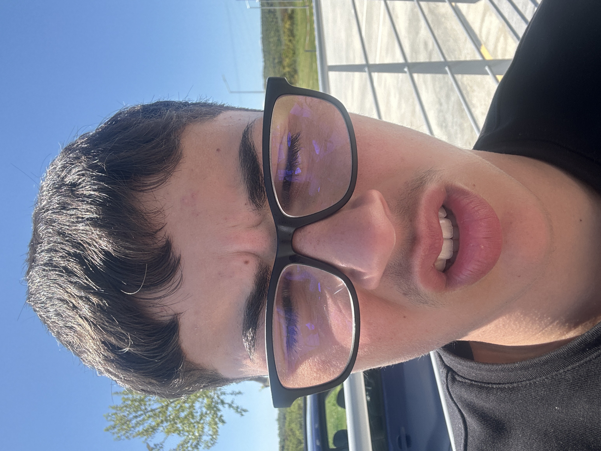 A close-up, slightly angled shot of a person wearing black-framed glasses. The person has dark hair, thick eyebrows, and fair skin. The glasses reflect the surrounding environment, showing hints of greenery and sky. Part of a black t-shirt is visible at the lower right. In the background, through the glasses' reflection, there's a glimpse of trees and a bright sky. The location in the city of Νέα Κερδύλια, Greece is assumed based on the prompt but cannot be confirmed visually from this image.
