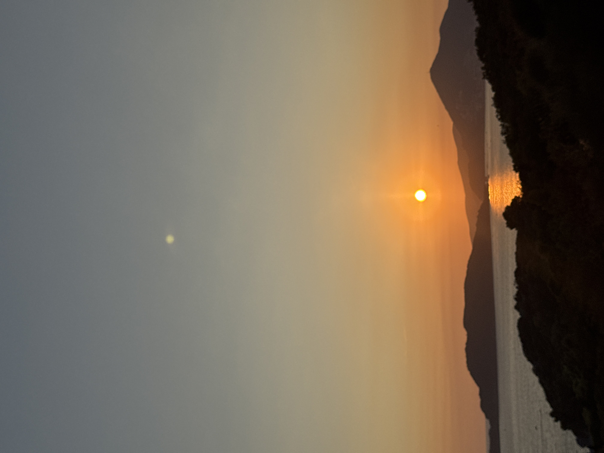 This is a vertical, outdoor, natural landscape photograph taken at sunset in Sveti Stefan, Montenegro. The image captures a serene coastal scene with a focus on the sky and water. The sky transitions from a pale blue at the top to a warm orange and yellow gradient near the horizon, indicating the setting sun. The sun, a bright orange orb, is positioned slightly left of center in the middle section of the image, casting a shimmering golden reflection on the water below.
In the foreground, dark, silhouetted hills covered in dense foliage create a textured foreground. Beyond these hills, a body of water, likely the Adriatic Sea, extends towards the horizon. Several islands or peninsulas, also appearing as dark silhouettes, are visible in the distance, framing the water. These landmasses have undulating shapes, with one prominent mountain peak standing out against the sunset. The overall atmosphere is peaceful and tranquil, characteristic of a sunset over a coastal region. There are no people or man-made structures immediately visible, emphasizing the natural beauty of the location. The image is slightly soft, possibly due to low light conditions or camera settings.
