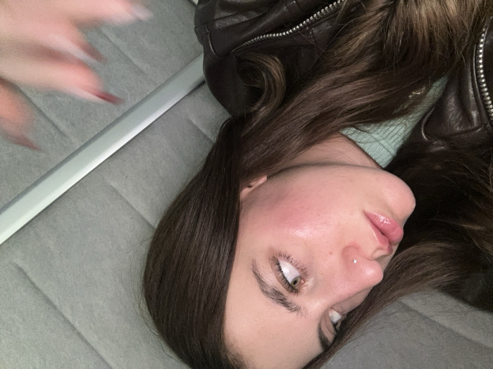 A woman with long, dark hair lies sideways on a grey upholstered surface, possibly a bed or couch. She wears a brown leather jacket over a light green sweater. Her skin appears dewy and she has a piercing on her nose. One hand with elongated, pointed nails is blurred in the upper left corner. The photograph is taken at an angle, making the composition slightly disorienting.