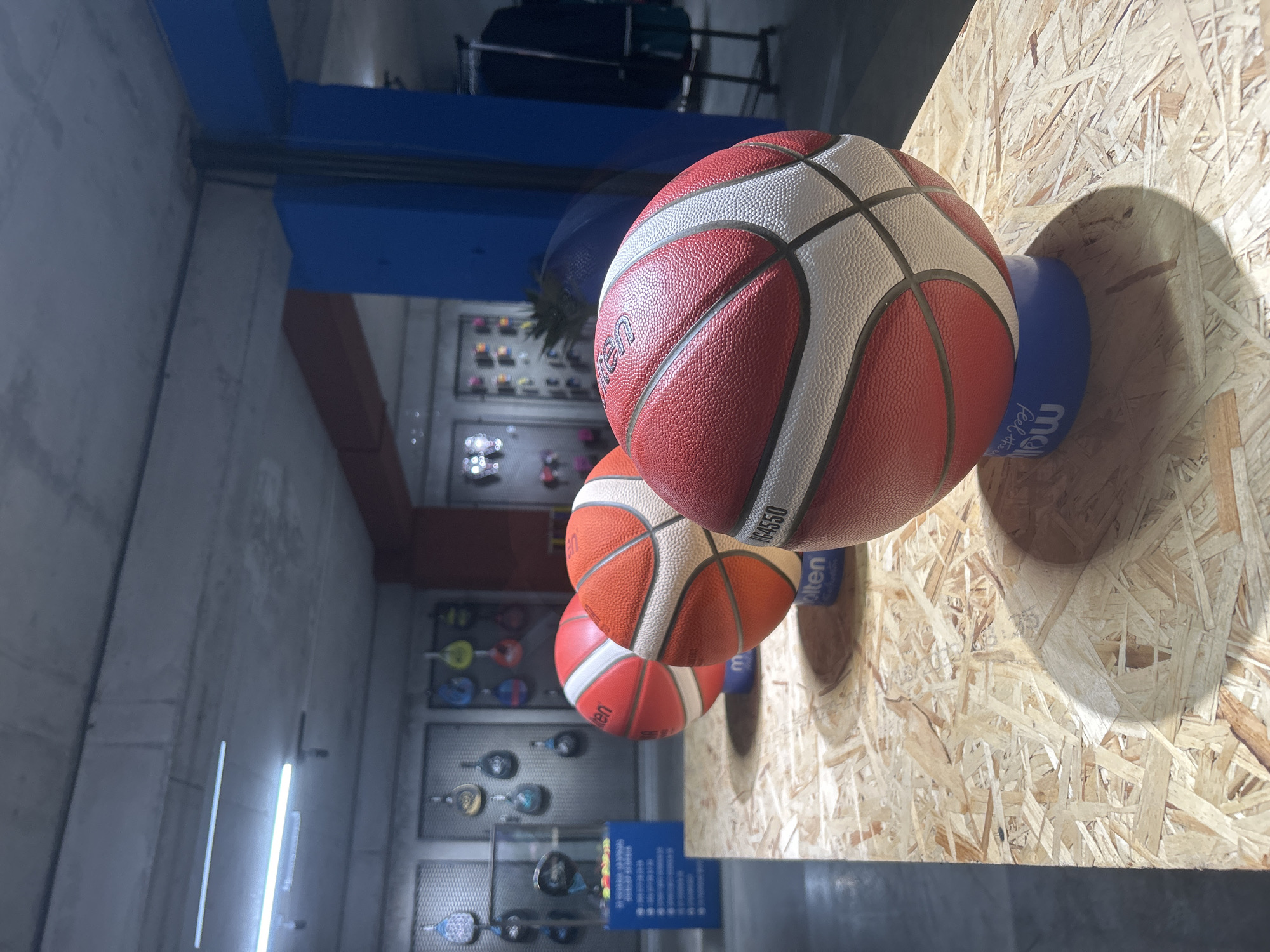The image shows a close-up of three basketballs on a wooden surface. The basketballs are red and white with black lines, and appear to be manufactured by Molten, as indicated by the branding on the balls. The basketball in the foreground is red and white, with the Molten logo and "GF750" printed on it. The other two basketballs are partially visible behind it, one of which is also red and white. The basketballs are resting on blue stands.
The background of the image is blurred but suggests an indoor retail environment, possibly a sports store. There are shelves with various sports equipment, including what appear to be paddles or rackets hanging on the wall. A blue pillar is visible on the right side of the image. The lighting suggests an indoor setting, possibly with fluorescent lights overhead.
The location is identified as Tbilisi, Georgia, specifically within a store called "Sportmaster" located in the East Point mall. This information comes from the text visible on a blue stand in the background which lists store details, including "Sportmaster Tbilisi" and a phone number that starts with +995 322 25 25 25.