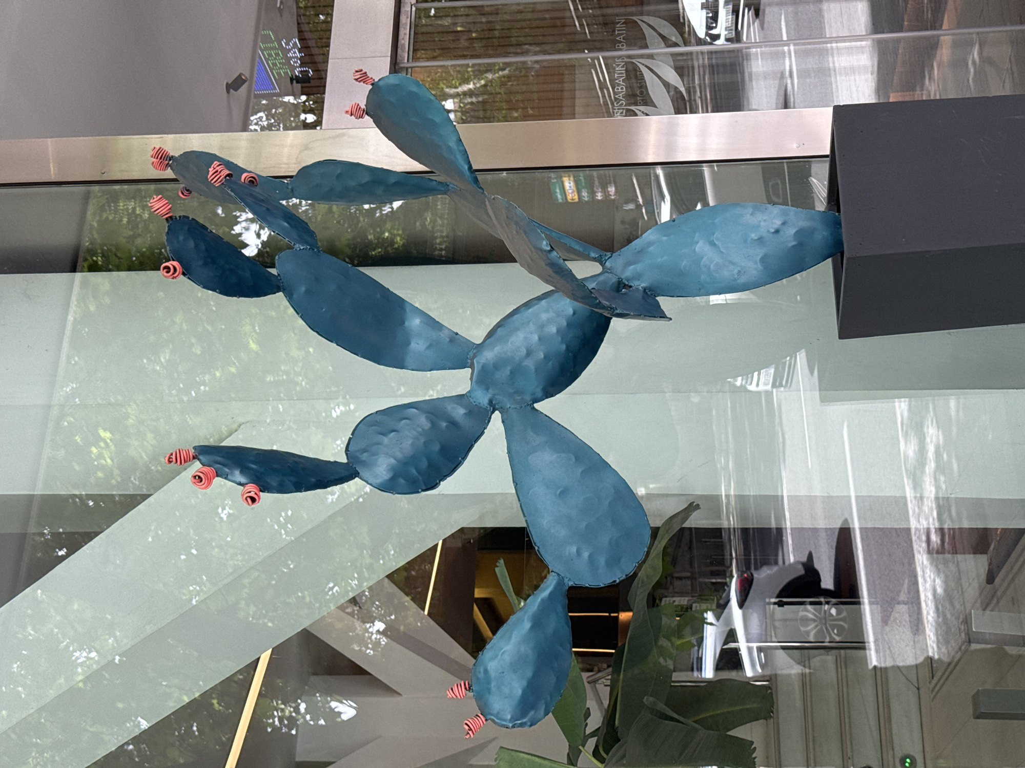 A blue metal prickly pear cactus sculpture is displayed in a window in Madrid, Spain. The sculpture has several pads with orange and red spines on the ends. The sculpture is mounted on a gray box. Reflections of trees and buildings are visible on the window. The lettering "NS SABATINS BATINI RTO SUIT" can be seen.