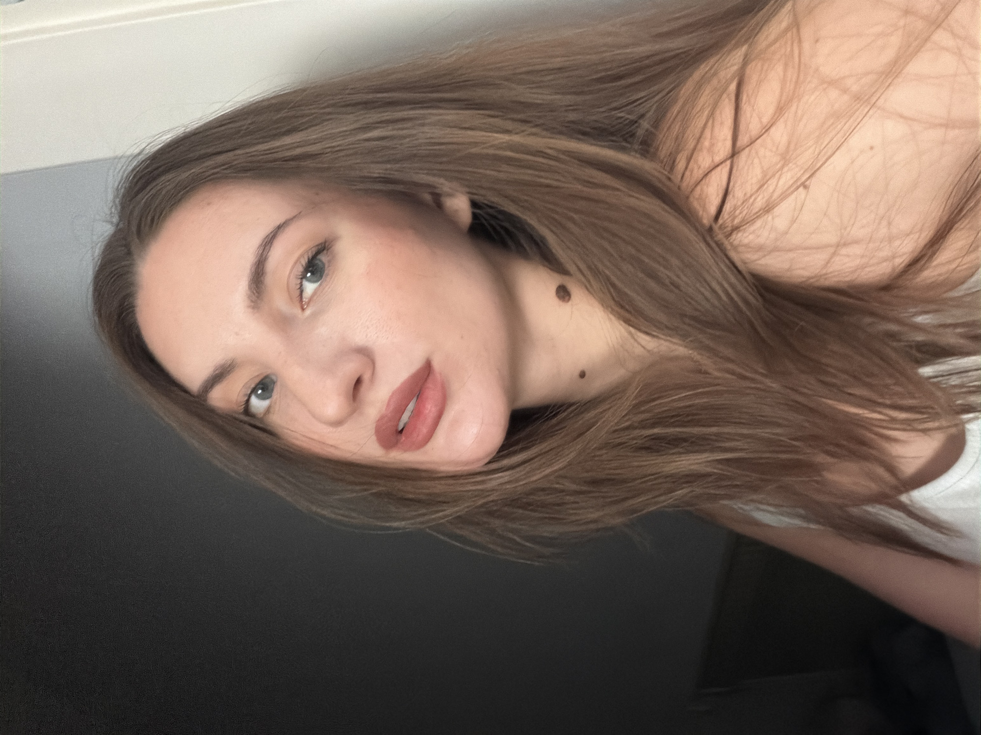 This media file is a close-up, angled portrait of a young woman. Her head is tilted, and her long, light brown hair cascades around her face and shoulder. She is looking directly at the camera with a neutral expression. Her skin appears fair with some subtle makeup, including blush and lipstick in a muted pink shade. A small, dark mole is visible on her neck.
The background is dark and out of focus, suggesting an indoor setting with low lighting. The top portion of the image shows a lighter, blurred area that could be a wall or ceiling.
There is no discernible activity or event taking place; it is a posed portrait. No text is visible in the image. The context of Sharm El Sheikh, Egypt, is provided but not visually evident from the image itself.