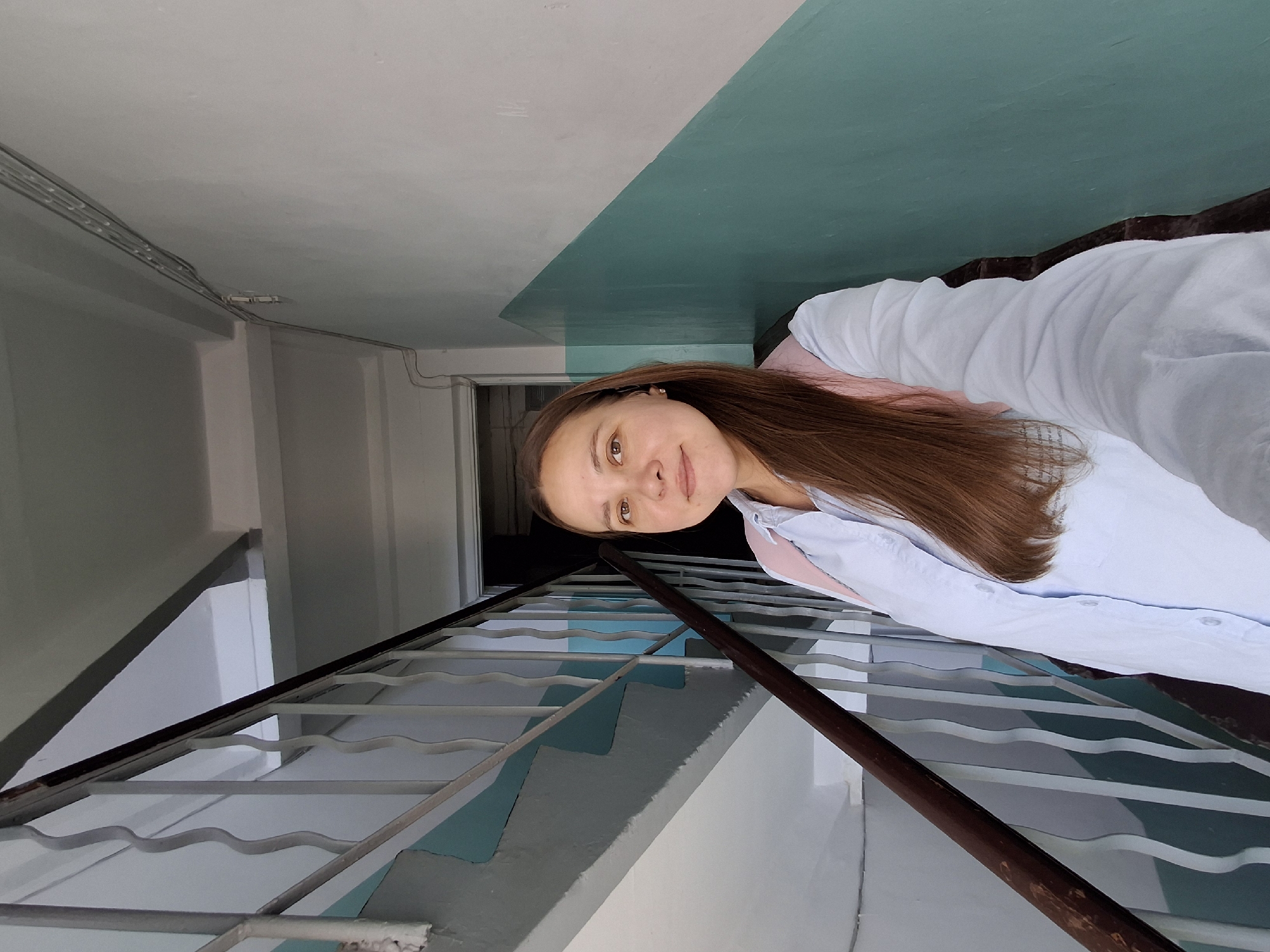 A young woman with long brown hair is taking a selfie in a dimly lit indoor stairwell. She is wearing a light blue button-down shirt and a pink backpack. The stairwell has white walls with a teal diagonal stripe, a wooden handrail, and metal railings. The lighting suggests it is daytime, but the interior nature of the stairwell makes it dark. The scene appears to be in a residential building, typical of Chaikovsky, Russia, given the style of the building's interior.