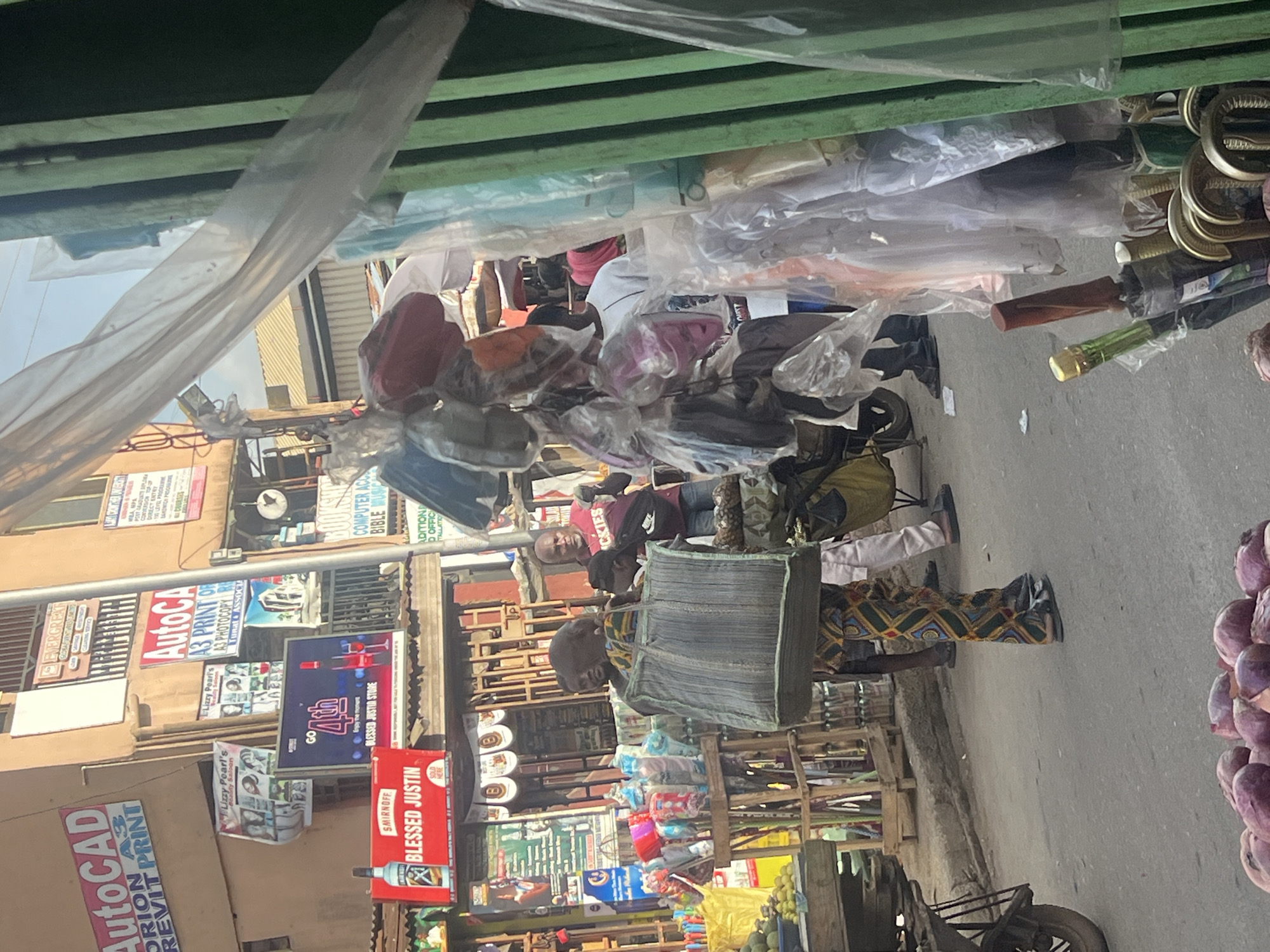 The image shows a street scene in Egan, Nigeria. Underneath a green awning, a person carrying a large grey bag is standing on a sidewalk. Several people are standing around them, including one wearing a red shirt and another in white. Street vendors display various items, including purses and umbrellas. Signs advertising "AutoCAD," "Blessed Justin," and "Lizzy Pearl's Beauty Saloon" are visible on the surrounding buildings. A pile of purple fruits is at the edge of the frame.