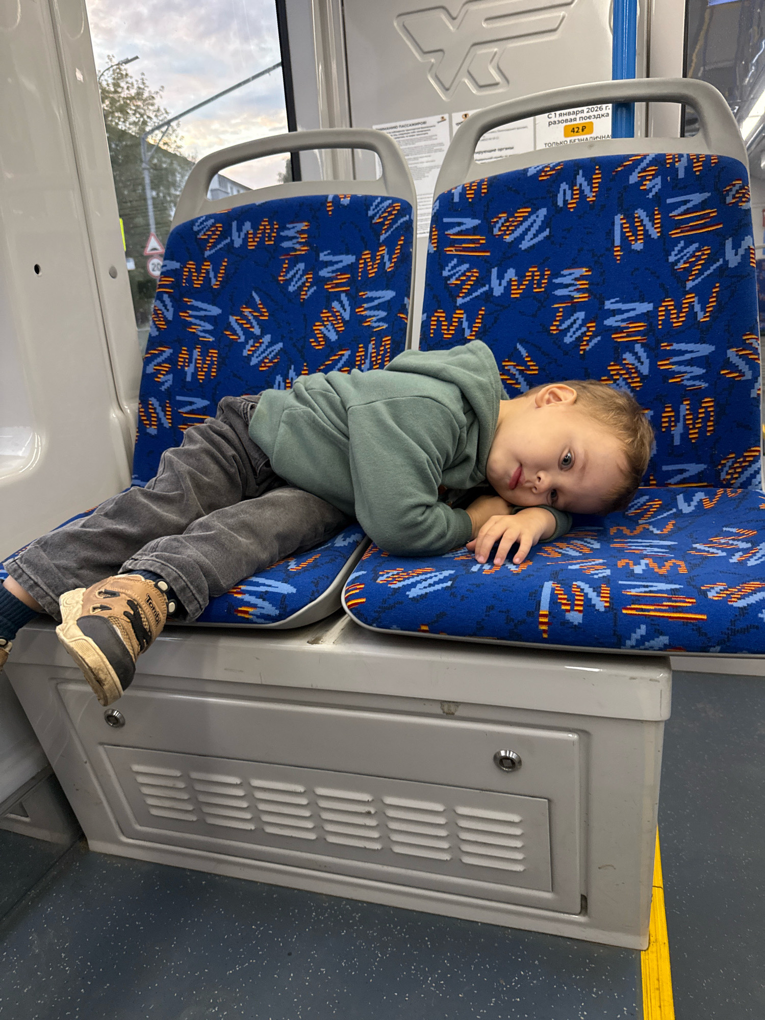A young Caucasian boy is lying on his side on a blue seat with a colorful pattern inside a public transport vehicle. The boy, with light brown hair and blue eyes, is wearing a light green hooded sweatshirt and dark grey pants. He is resting his head on his hands, looking towards the camera with a slightly pensive expression. His feet, wearing brown and black shoes, are visible on the seat. The seats in the vehicle are upholstered in a blue fabric with a repeating abstract pattern of orange and blue shapes. A sign with Russian text and a price is visible above the seat. The environment outside the window shows trees and a cloudy sky. The floor of the vehicle is dark grey with small flecks of white. The overall scene appears to be within the city of Rusanovo, Russia.