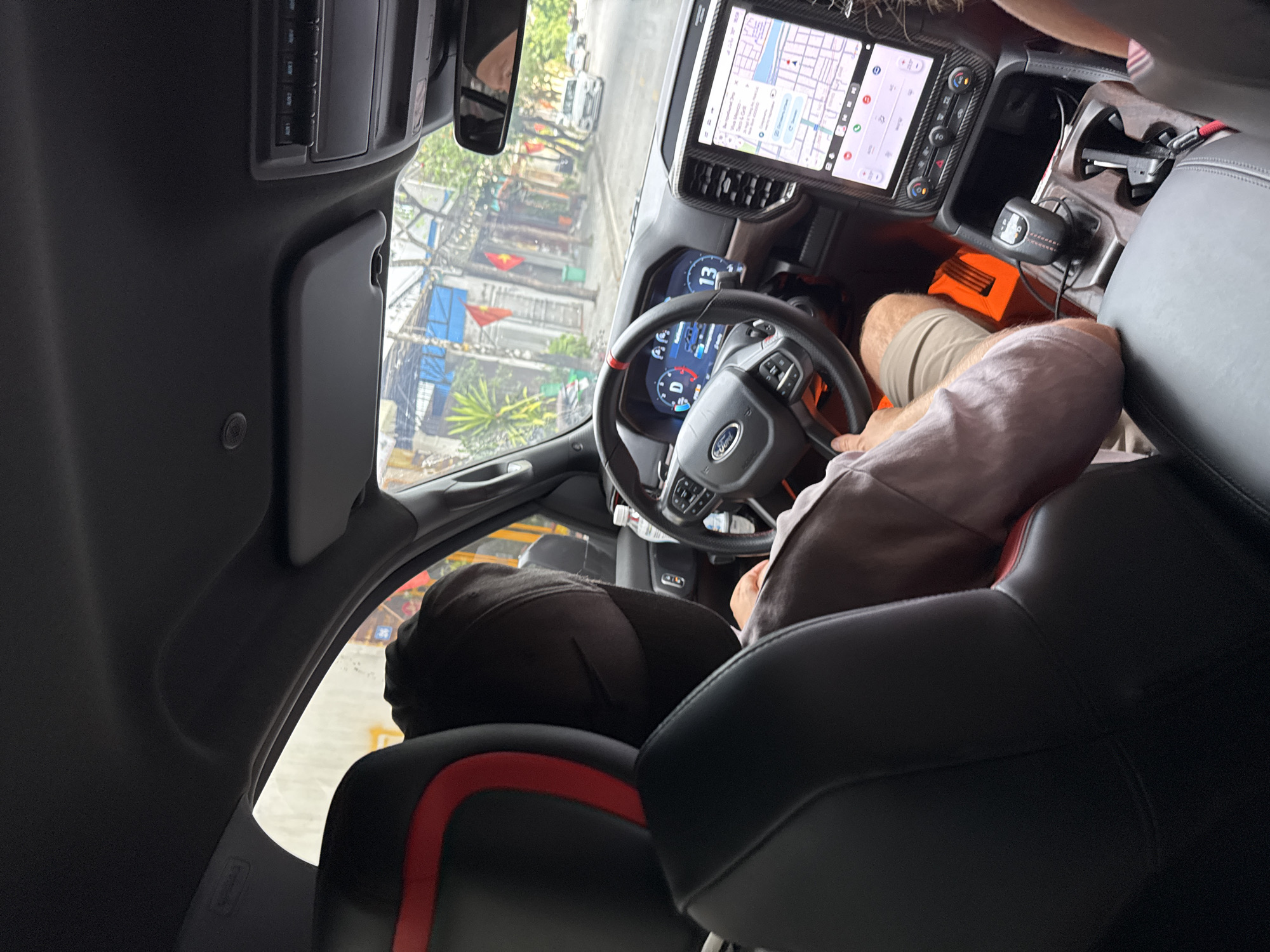 The image shows the interior of a Ford vehicle, viewed from the rear passenger seat, looking forward. The driver, wearing a black face mask and a light-colored shirt, is operating the vehicle. The dashboard features a digital instrument cluster displaying "D" for drive and a speed of 13, along with a large central touchscreen. The exterior view through the windshield reveals a street scene in what is identified as Hoa Lư Ward, Vietnam. Buildings with balconies and trees line the street, and Vietnamese flags are visible, indicating the location. The interior of the car is modern, with leather seats and ambient lighting. A bottle of water is visible on the dashboard. The overall scene depicts a person driving a vehicle in a city environment in Vietnam.