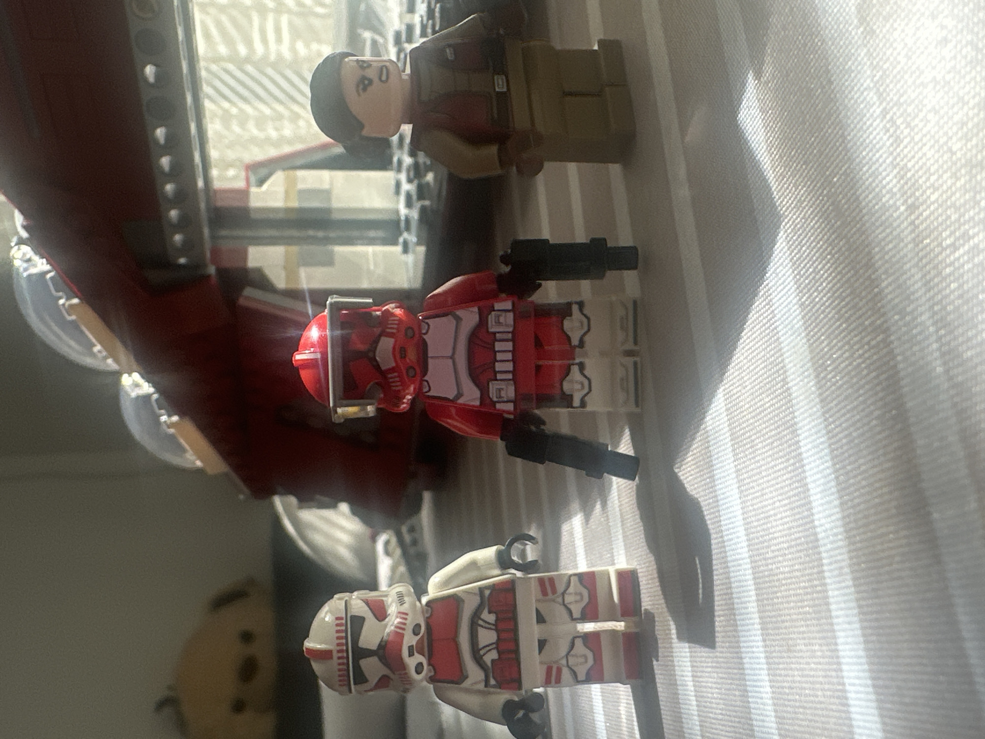 Three Lego minifigures are the primary subjects of this image, arranged on a light-colored, textured surface, possibly a window sill or table. On the far left, part of a large red Lego structure, likely a vehicle or spacecraft, is partially visible.
Starting from the top of the image (which is oriented vertically), the first minifigure is a human-like character with dark hair, a light skin tone, and is wearing a brown and tan outfit. Below this figure, a red and white Lego clone trooper or stormtrooper stands, holding two black blaster pistols. Further down, a second, similarly styled red and white Lego clone trooper or stormtrooper lies on its back, displaying its patterned armor. In the blurred background on the far left, a soft, tan-colored object with dark features, possibly another toy or plushie, is faintly visible.
The scene is set indoors, against a backdrop of bright, vertically striped, textured fabric, resembling window blinds or curtains. Strong, likely natural light, illuminates the scene from the left, casting distinct shadows of the minifigures and the red structure onto the background fabric. The overall impression is a display or play setup of Lego figures.
There is no discernible activity taking place beyond the static arrangement of the figures. The bright lighting suggests it is daytime. No specific location cues for Sofia, Bulgaria, are visible within the image itself. No text is visible in the media file.