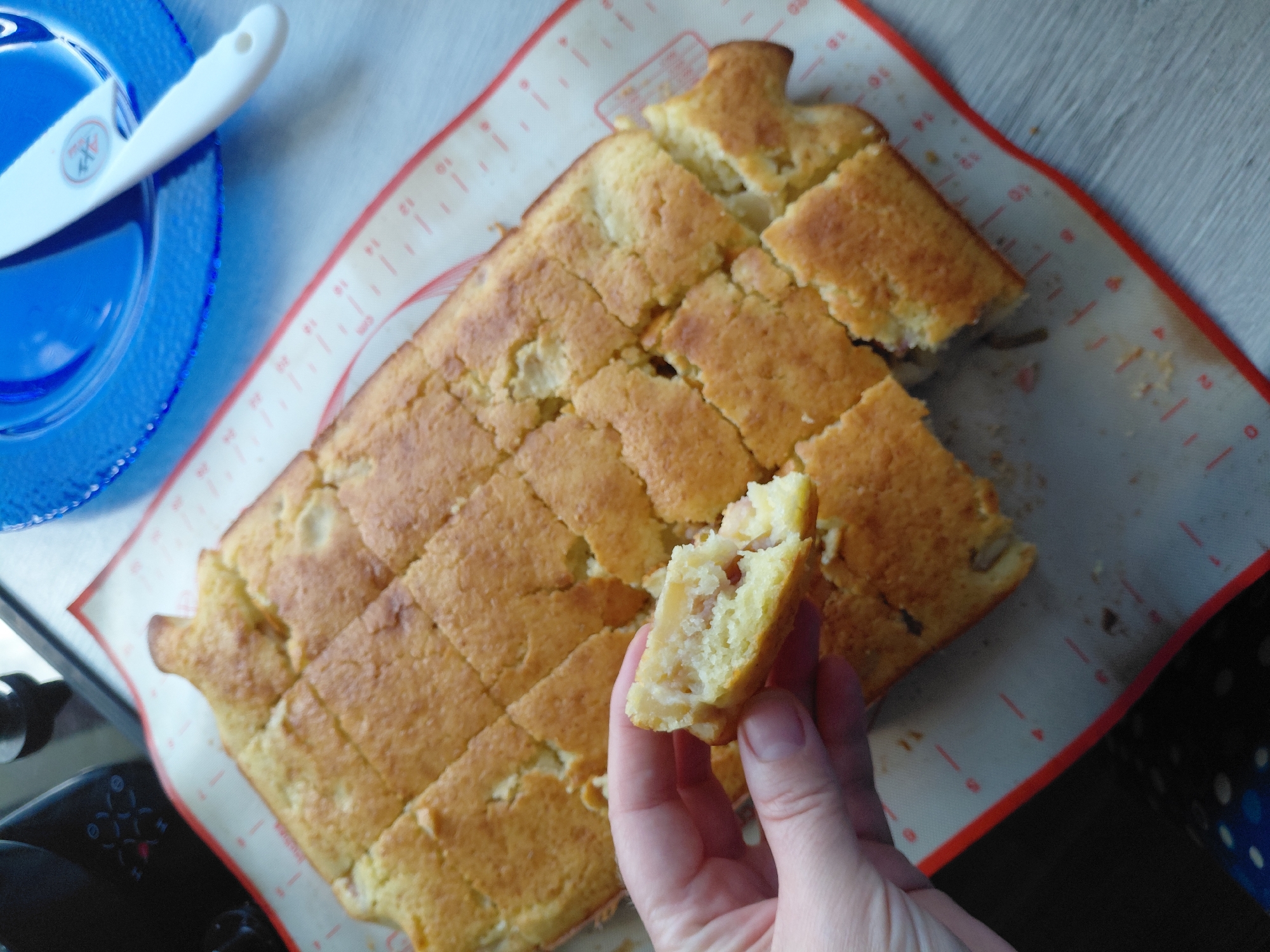 A hand holds a piece of a baked good, which has been cut into a grid of squares. The baked good appears to be a cake or bread, with a golden-brown crust and a lighter interior that contains visible pieces of fruit or nuts. It is resting on a red-edged baking mat with grid lines, placed on a light-colored wooden surface. In the background, a blue plate with a white utensil and a black kitchen appliance are partially visible. The scene is indoors, likely a kitchen. There is no indication of the specific location in Novosibirsk, Russia.