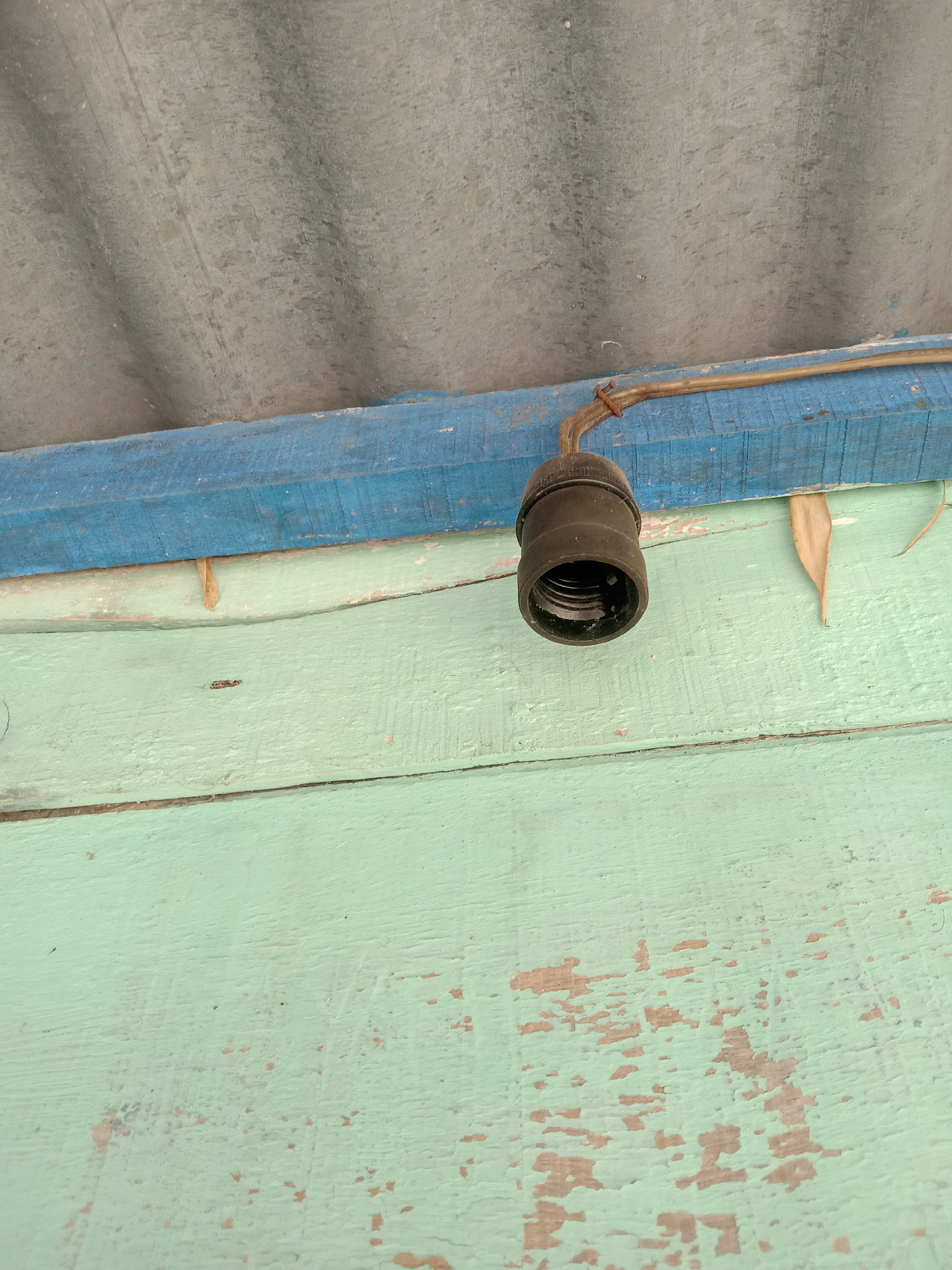 An outdoor image depicts a close-up view of a light socket hanging from wires attached to a blue wooden beam. The light socket is black and appears to be empty. Above the blue beam is a corrugated, grey roof. Below the blue beam is a light green wooden surface, possibly a wall or fence, showing signs of wear with chipped paint. A dry, brown leaf is visible near the light socket, caught between the blue beam and the green surface. The image captures a scene that could be part of the exterior of a building or structure in Stiaji, Indonesia.