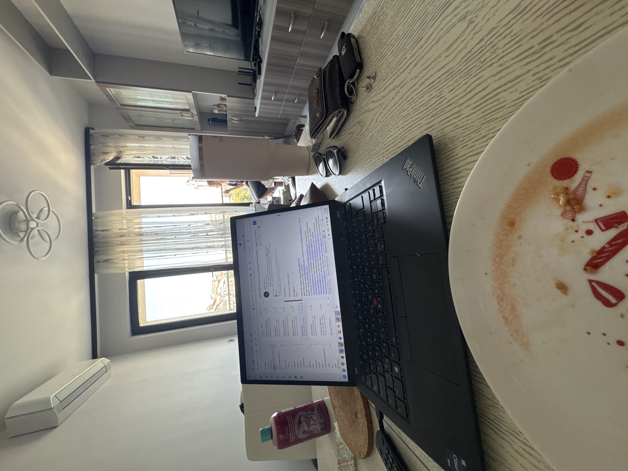 The image depicts an indoor scene, likely a home office or a studio apartment, with a table in the foreground holding a laptop, a dirty plate with remnants of food, a bottle of pink liquid, sunglasses, a wallet, and keys. The laptop screen displays an email inbox. In the background, a person is visible near an open balcony door, and an air conditioning unit is mounted on the wall. Furnishings suggest a living space with shelves and a television. The ambient light suggests daytime. Without further context or discernible landmarks, pinpointing the exact location within Sofia, Bulgaria is not possible from the visual information alone.