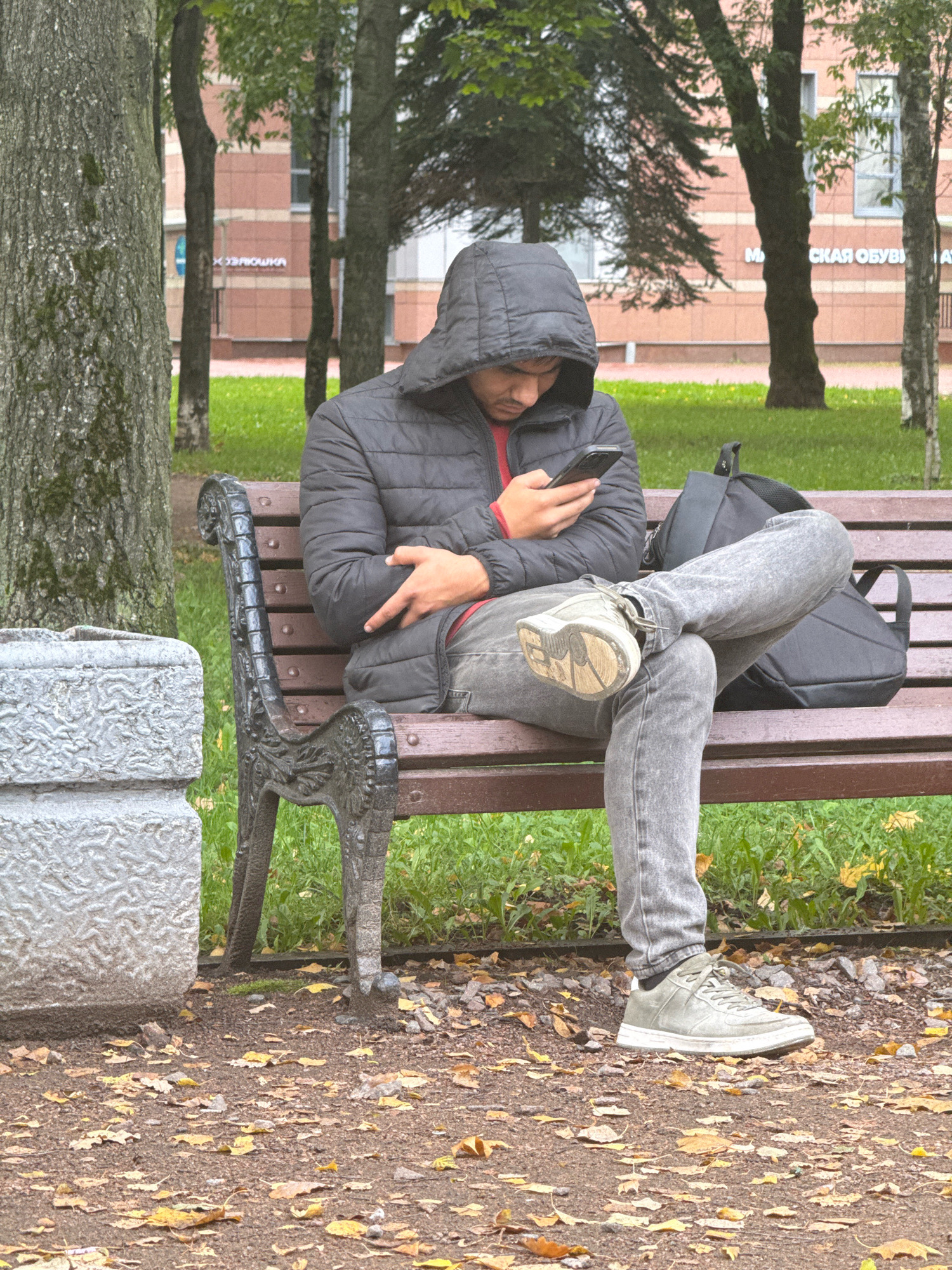 A young man, wearing a hooded jacket and grey jeans, sits on a park bench with his legs crossed, looking at his mobile phone. A black backpack rests on the bench beside him. The scene is outdoors, with trees and buildings visible in the background. The ground is covered with fallen autumn leaves, suggesting a park or a tree-lined street in Saint Petersburg, Russia. The bench appears to be made of wood and cast iron.
