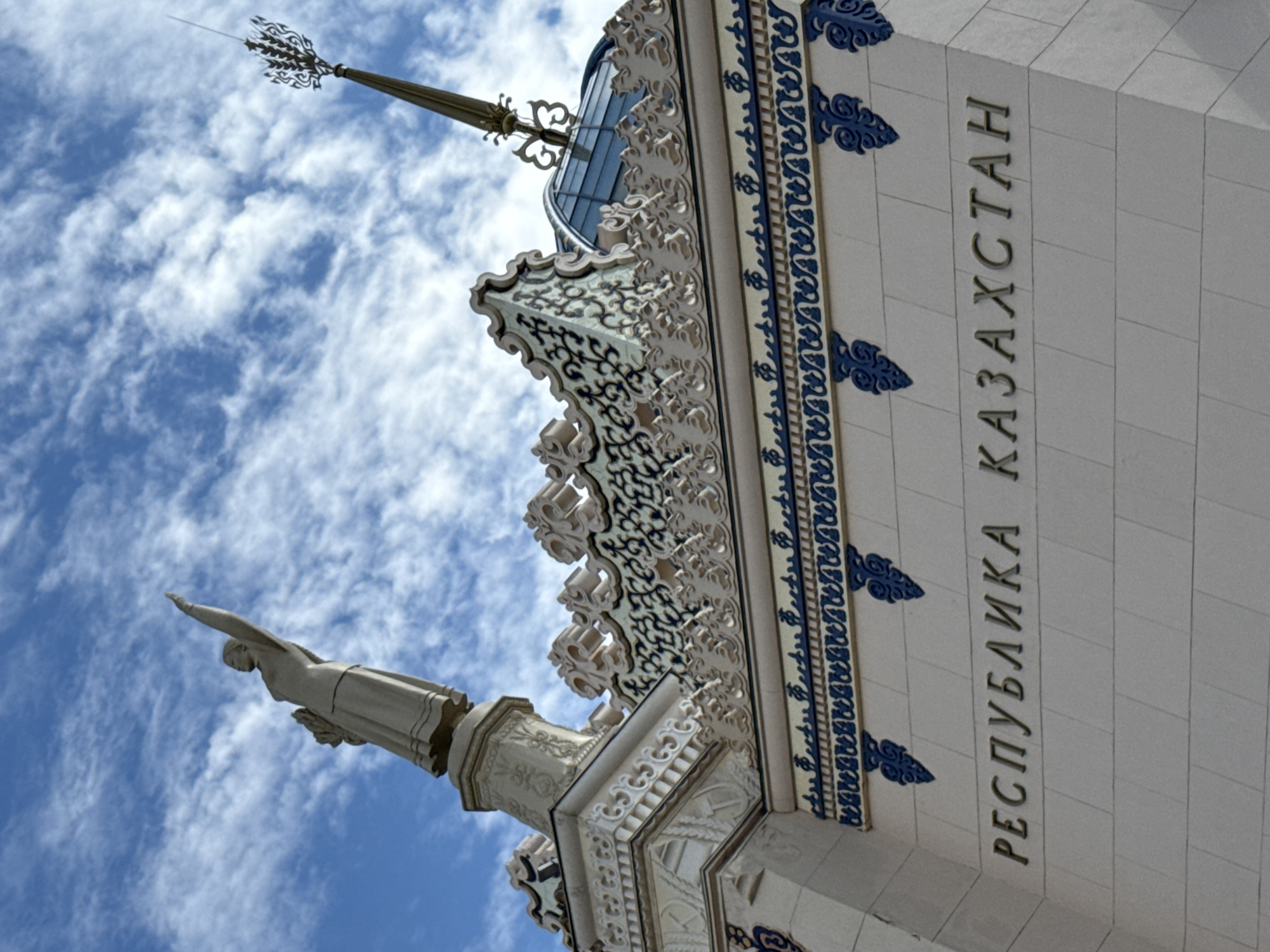 A low-angle, wide shot captures a section of an ornate building against a bright blue sky filled with scattered white clouds, suggesting a clear and sunny day. The building's facade features intricate decorative elements, including elaborate moldings and blue ornamental patterns. Prominently displayed on the building are large, metallic letters spelling out "РЕСПУБЛИКА КАЗАХСТАН" vertically, indicating that this is the pavilion or a building associated with Kazakhstan.
To the left of the text, a portion of a white stone statue of a woman with outstretched arms is visible. The statue appears to be in motion, possibly representing victory or aspiration. Above the statue and to the left, a decorative spire rises against the sky, adding to the grandeur of the architecture. The overall scene depicts an impressive architectural structure, likely a landmark or part of an exhibition, in Moscow, Russia. The lighting and sky suggest it is daytime.