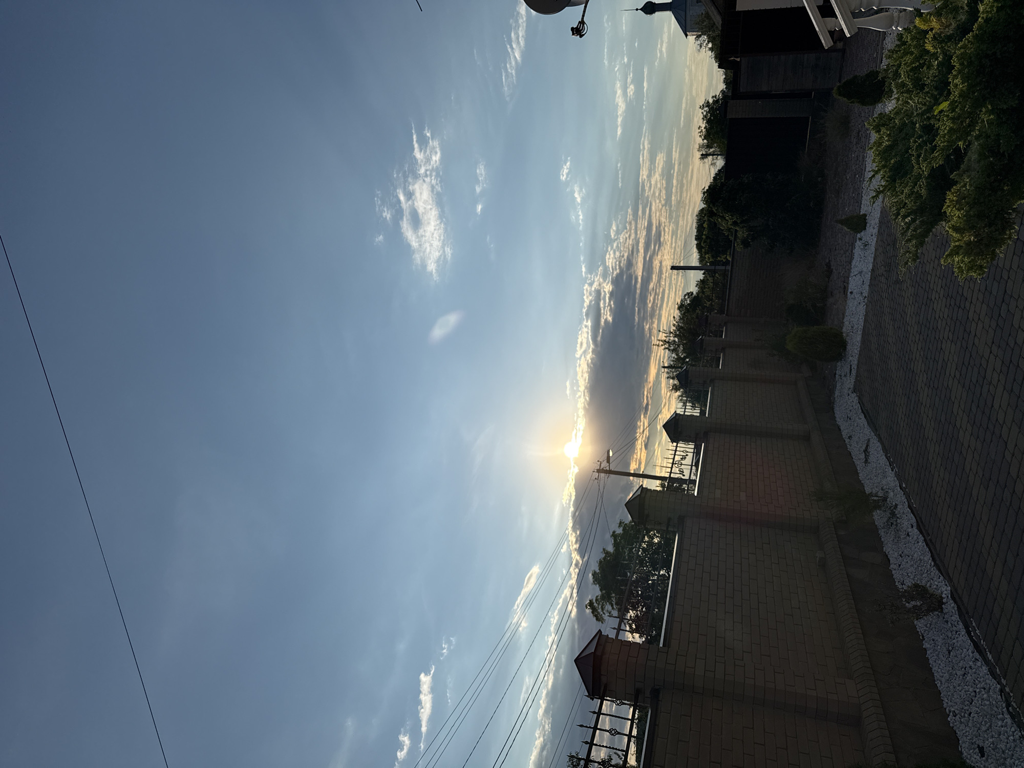 The image shows a brick fence with decorative metal elements and stone bases in Horbanivka, Ukraine, at sunset. The fence encloses a paved area with white gravel and small, manicured shrubs. In the background, trees and a church steeple are visible. The sky is a pale blue with scattered clouds, and the sun is setting behind a bank of clouds, casting a warm glow. Overhead, power lines are strung across the sky.