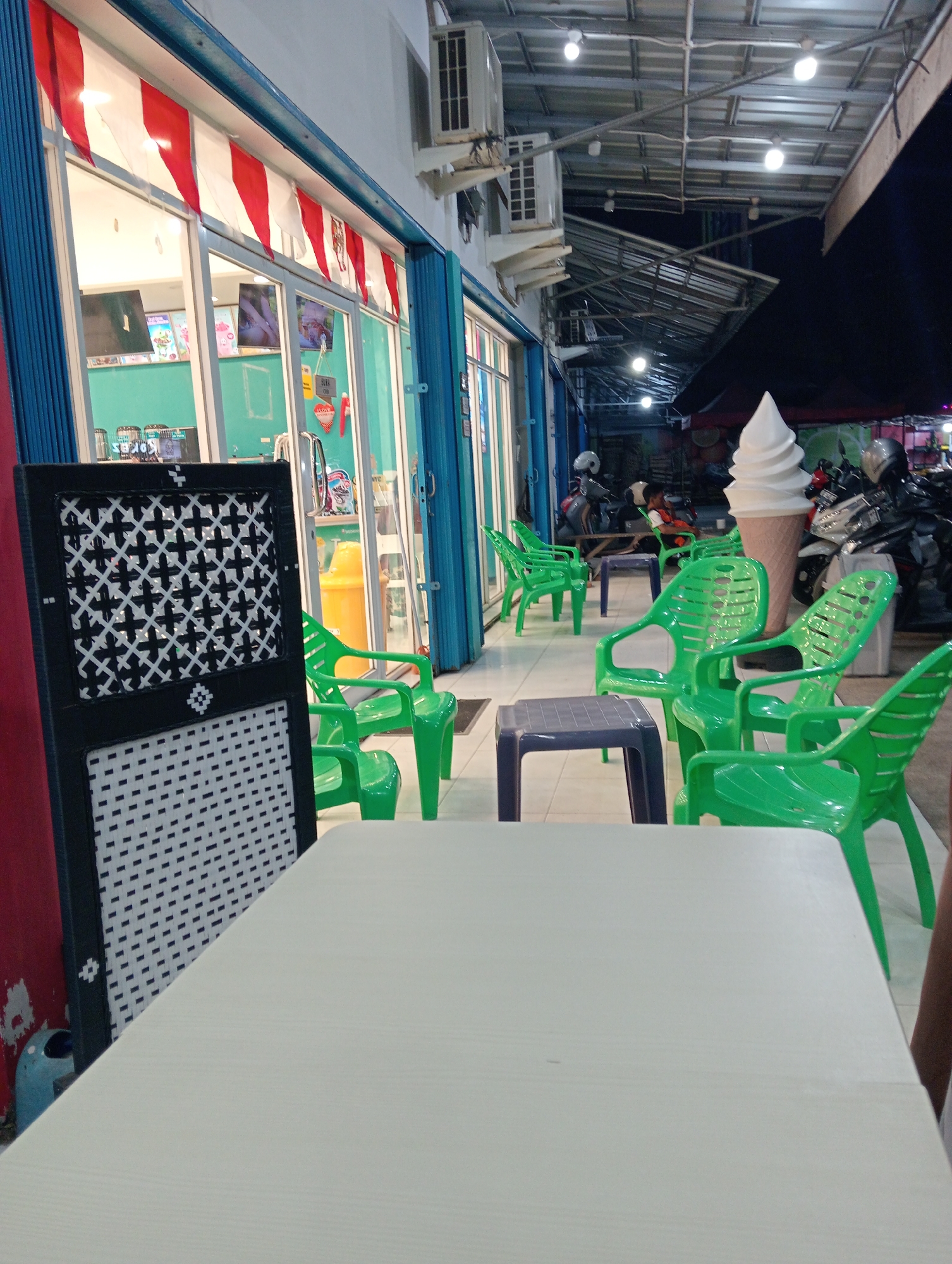 The image depicts an outdoor seating area of a shop, likely an ice cream parlor, in Pringsewu, Indonesia. The shop front, characterized by a blue collapsible door and large glass windows displaying ice cream products, is adorned with red and white triangular flags. Outside, a row of bright green plastic chairs and dark blue plastic tables are arranged on a tiled floor. In the background, several motorcycles are parked under a covered walkway, illuminated by overhead lights. A large, cartoonish ice cream cone sculpture stands prominently near the motorcycles. The scene appears to be captured in the evening, with ambient light sources illuminating the area.