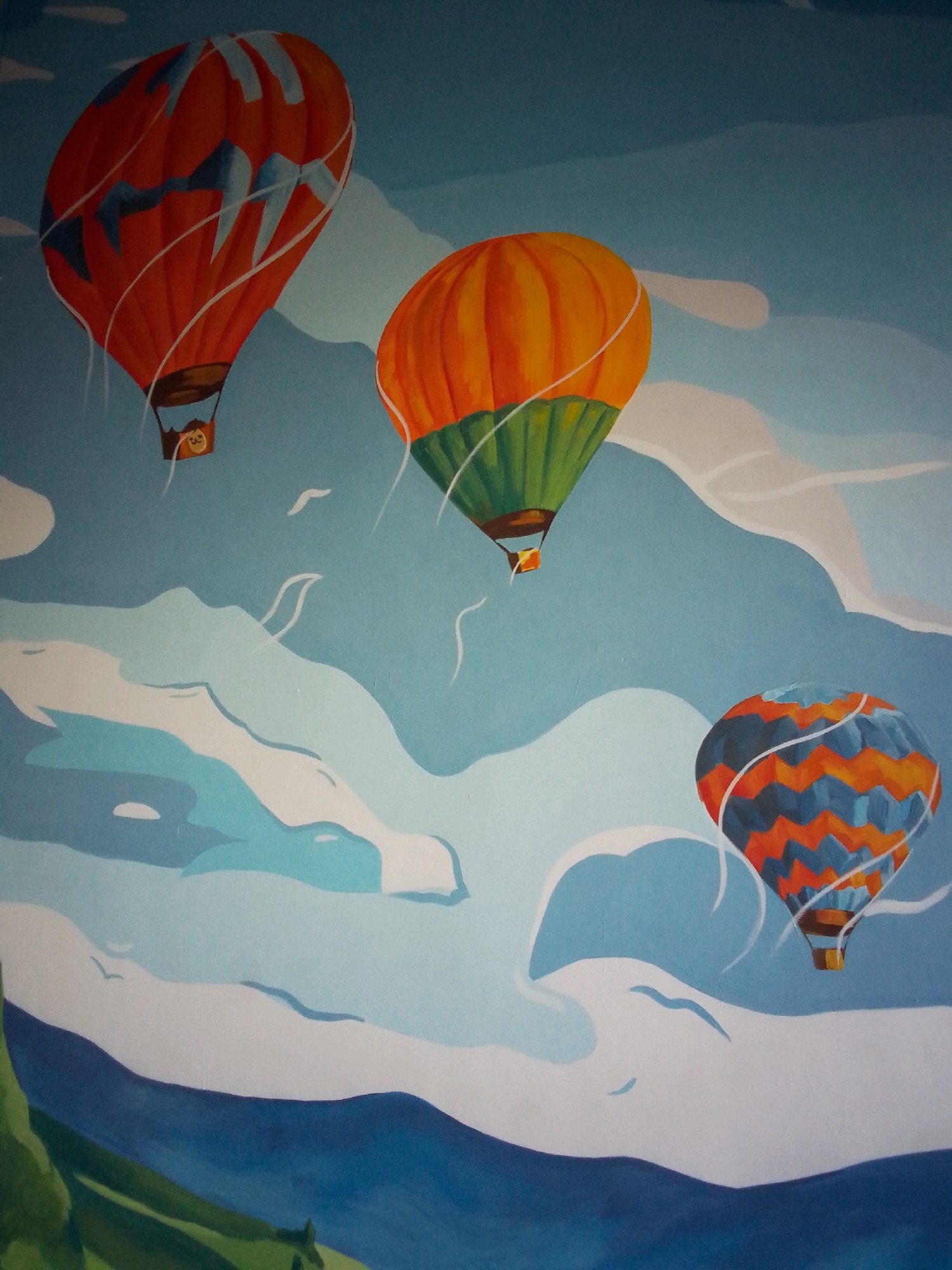 This is a painting of three hot air balloons floating in a blue sky with stylized white clouds and a distant blue mountain range. The hot air balloons are depicted in a cartoonish, vibrant style. The balloon on the left is orange with light blue markings and a brown basket. The middle balloon is orange on top and green on the bottom, also with a basket. The balloon on the right is a pattern of orange and blue zigzags, with a brown basket. The painting appears to be a mural or decoration on a wall, as evidenced by the textured surface and the way the painting blends into the surrounding environment, suggesting it is on an interior wall. It is not possible to determine the specific location in Tbilisi, Georgia from the image alone.