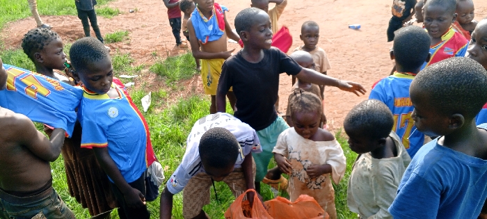 The image depicts an outdoor scene in Ngalula, Democratic Republic of the Congo. A group of approximately fifteen to twenty children, appearing to range from toddlers to pre-teens, is present on a surface composed of reddish-brown dirt interspersed with green grass patches. In the central foreground, a male child wearing a white t-shirt with a dark emblem and patterned brown shorts is observed bending over, focusing on an orange plastic bag situated on the ground. Adjacent to him, a smaller child in a light-colored, soiled garment also directs attention downwards. To the left, a male child, partially visible, holds a blue fabric item marked with the number '14' in yellow. In the mid-ground, a male child in a black t-shirt and light-colored shorts stands with his mouth open and right arm extended in a gesturing motion towards the right. Multiple other children, dressed in various t-shirts and shorts, are distributed throughout the frame, observing the central activity or each other. The background consists of exposed dirt and sparse vegetation.