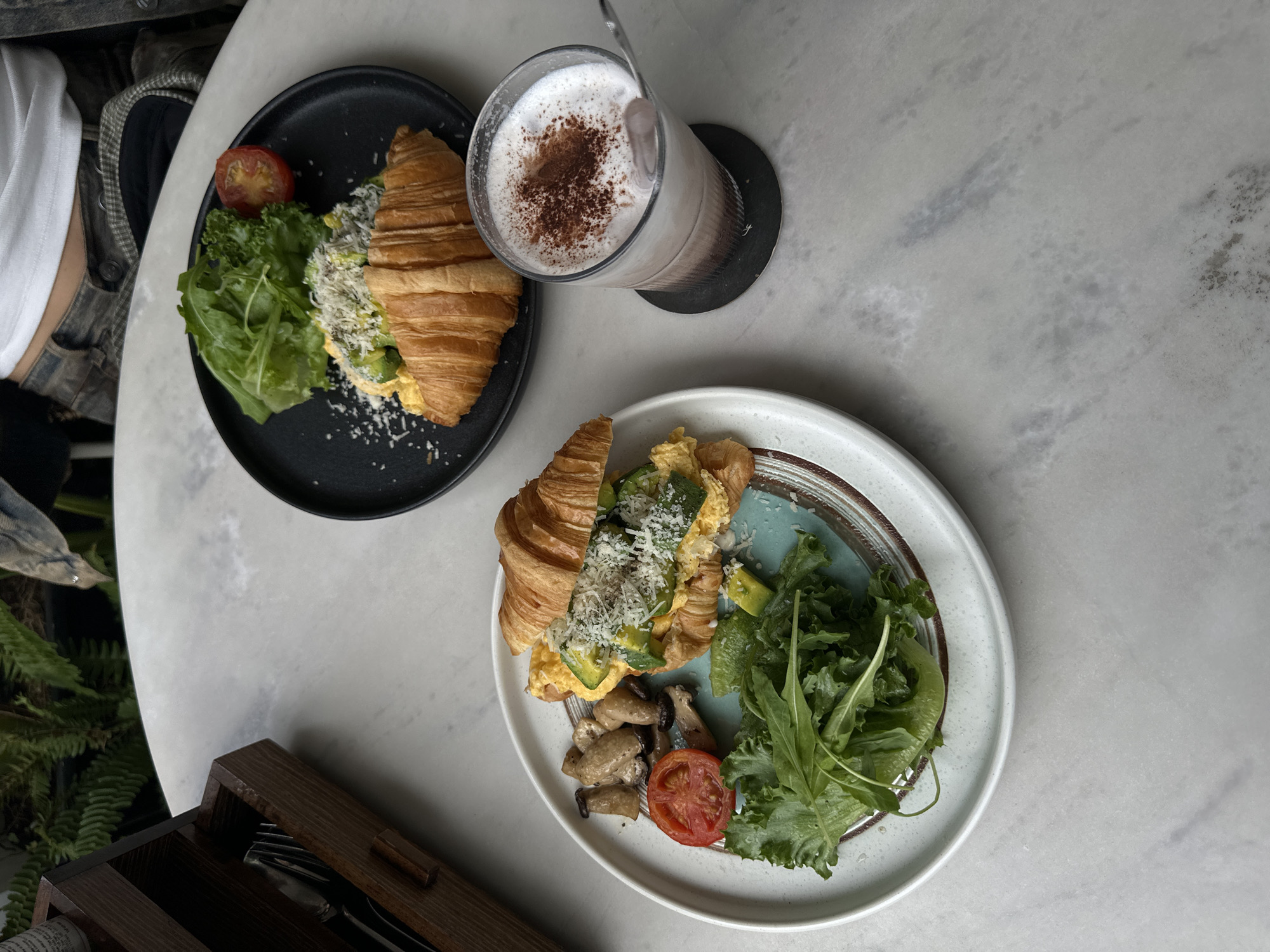 Two plates of croissant sandwiches with scrambled eggs, avocado, cheese, and greens are on a marble table. One plate also includes sliced tomatoes and mushrooms. A glass of iced coffee with chocolate powder sprinkled on top sits on a coaster. A wooden cutlery holder is to the left of the plates. The background shows greenery and a person's midsection wearing patterned pants and a white top. The scene suggests a breakfast or brunch setting in Da Lat, Vietnam.