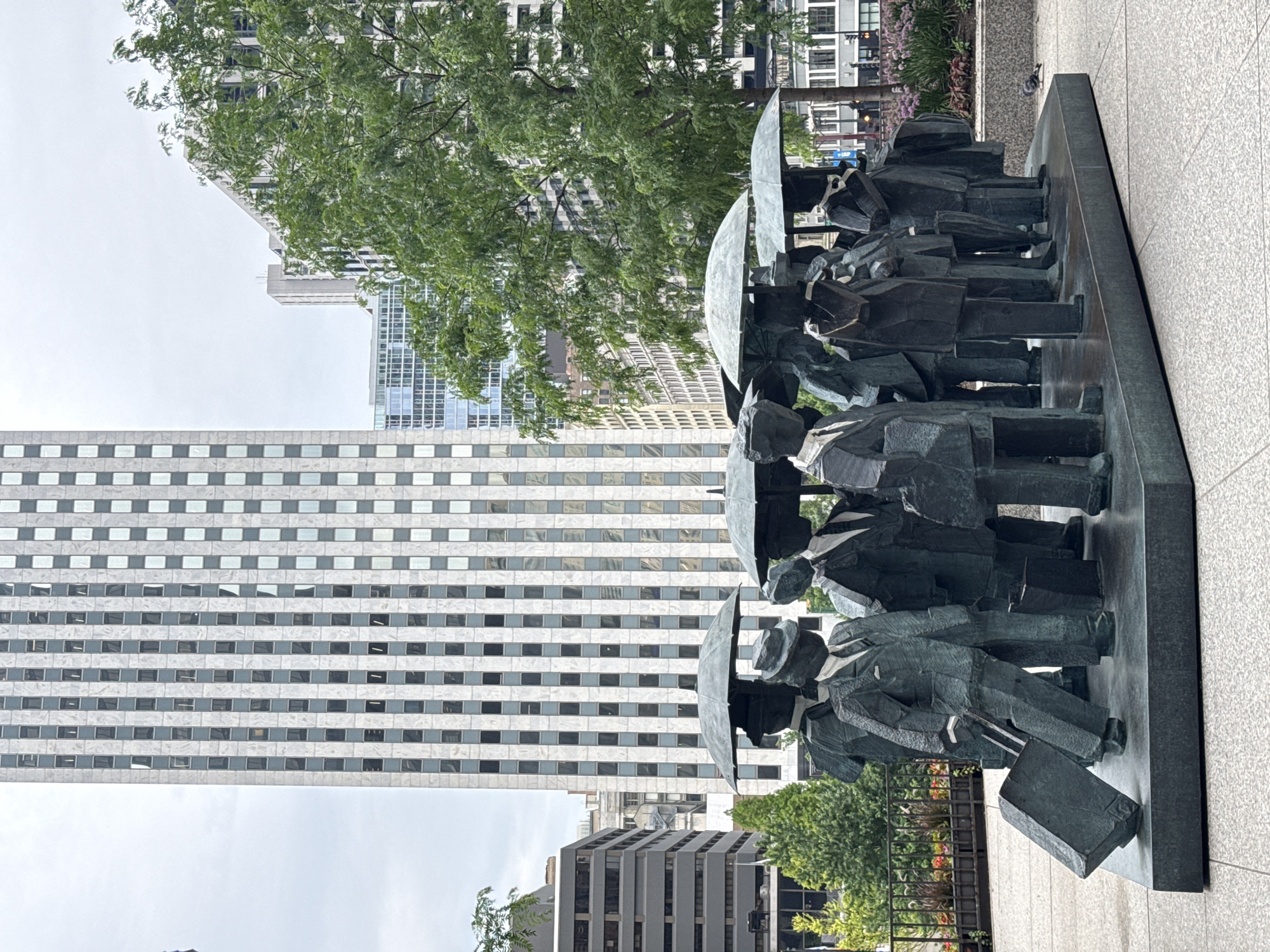 This media file features a bronze sculpture of a group of people standing in a line, each holding an umbrella and some carrying briefcases. The figures are stylized and abstract, with sharp angles and geometric shapes. They appear to be dressed in formal attire, suggesting they might be commuters or business people. The sculpture is positioned outdoors, with a backdrop of modern skyscrapers and greenery.
The activity depicted is ambiguous but could represent people waiting, commuting, or simply gathered together. The umbrellas suggest it might be raining or the sun is strong.
Notable details include the textured granite paving stones surrounding the sculpture, the numerous windows on the buildings, and the lush green trees. The lighting is overcast, indicating a cloudy day, likely mid-day. The overall mood of the scene is somewhat somber and stoic, conveyed by the rigid postures of the figures and the muted colors of the environment. The location is identifiable as Chicago, USA, due to the architectural style of the buildings.
There is no visible text in the image.