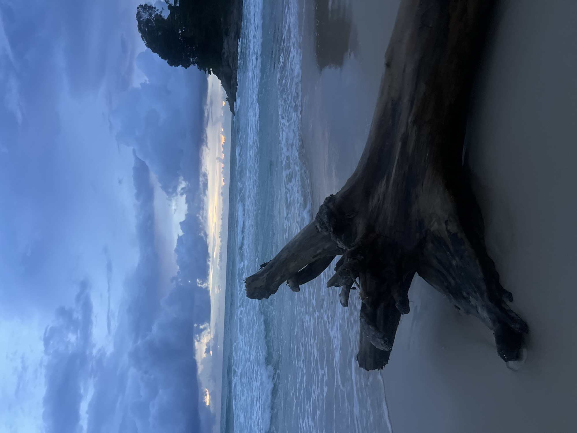 The image depicts a tranquil beach scene at dusk. In the foreground, a large piece of driftwood rests on the sand, its gnarled branches extending towards the ocean. The waves gently lap the shore, creating a soft, rhythmic pattern on the wet sand. The ocean itself appears calm, with subtle reflections of the sky on its surface.
The sky is filled with dramatic, brooding clouds in shades of deep blue and gray, interspersed with soft, warm light peeking through. This suggests the transition from day to night, possibly after a storm or as the sun sets. A faint sliver of orange light can be seen on the horizon, hinting at the last rays of sunlight.
To the left of the frame, a dark, tree-covered cliff juts out into the scene, providing a sense of scale and context to the vastness of the ocean and sky. The overall mood of the image is peaceful and contemplative, with a touch of dramatic beauty from the cloudy sky. The location is identified as Choeng Thale, Thailand. There is no visible text in the image.